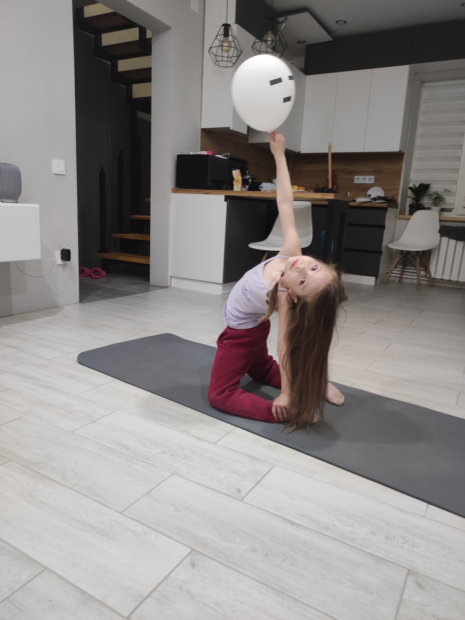 A young girl is performing a yoga pose on a gray mat indoors. She's kneeling with her torso twisted to the side, one arm raised and holding a white balloon with black tape forming a crude face. She is wearing a light-colored shirt and dark red pants. The background includes a kitchen area with white cabinets and a black countertop. There is also a staircase visible in the background. The location appears to be a home interior, possibly in Votkinsk, Russia, based on the prompt, but without visual confirmation of landmarks, this cannot be definitively stated.