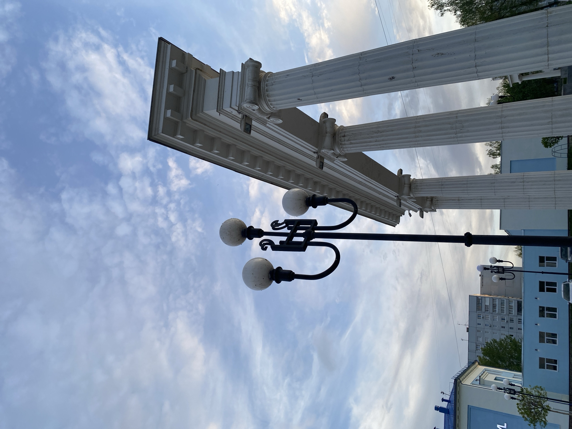 The image shows a view looking up at a classical architectural structure in Chaikovsky, Russia. Three white, fluted columns with ornate capitals support a white entablature. A black, three-globed lamppost is situated in front of the columns. The sky is visible behind the structure, filled with scattered clouds. In the background, buildings typical of an urban environment can be seen.