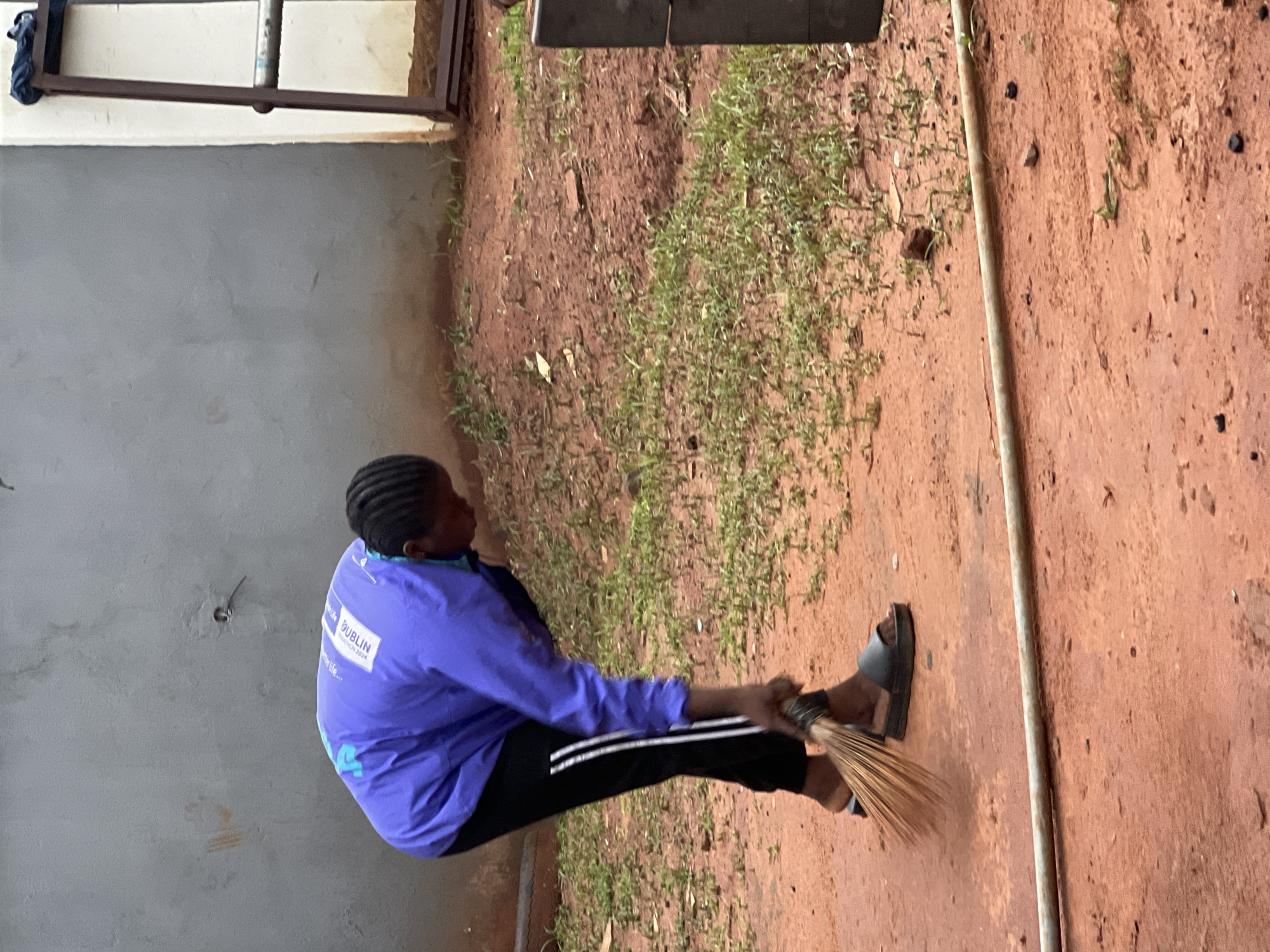 An individual is sweeping a dirt ground with a broom in Nkpor, Nigeria. The person is wearing a purple jacket with white lettering and black track pants with white stripes. They are also wearing black slide sandals. The ground is composed of reddish-brown dirt with sparse green vegetation. In the background, there is a gray wall and a metal frame structure with a glass pane. A metal pipe lies horizontally across the foreground.