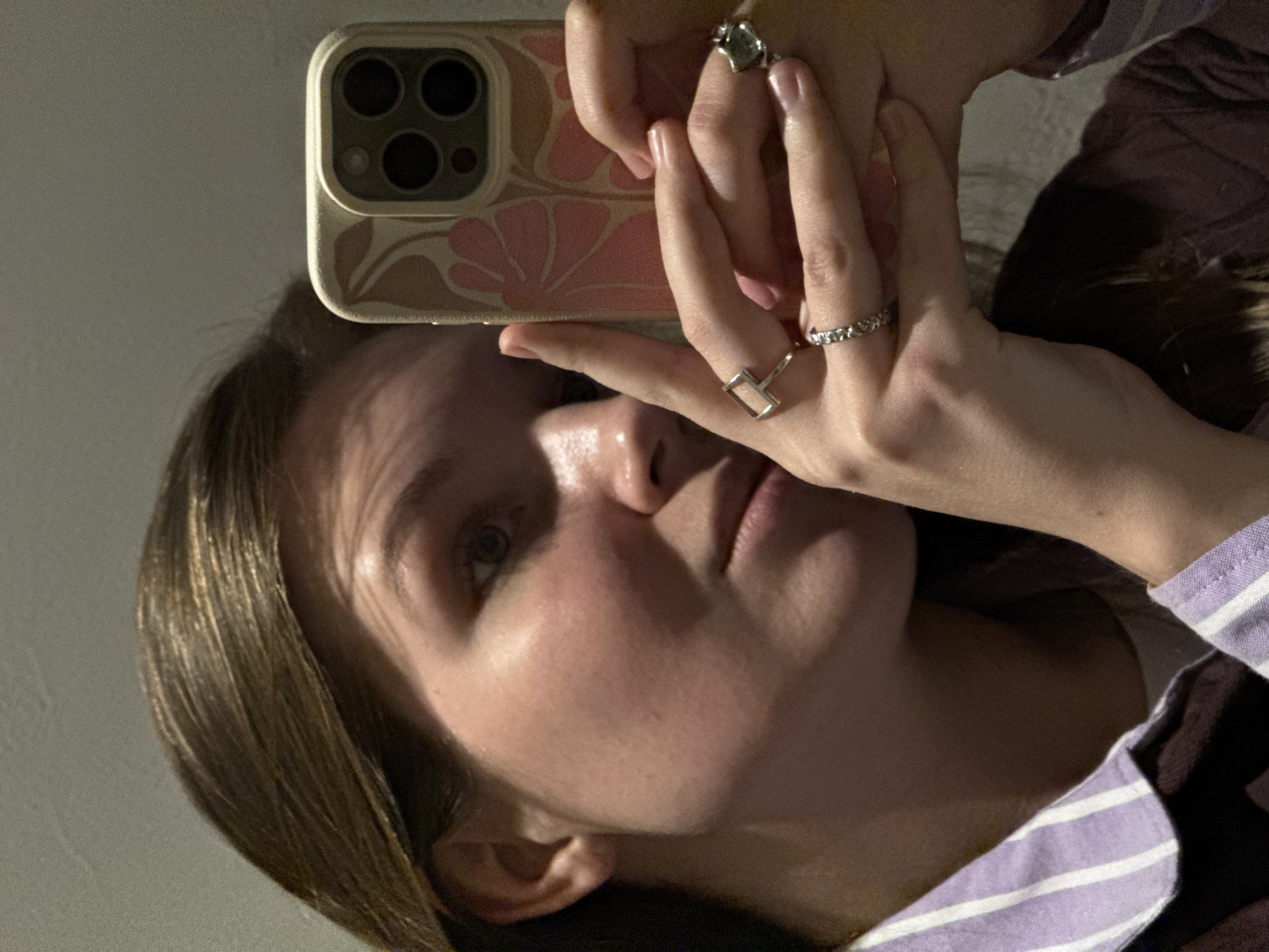 A young female, likely Caucasian, with medium-brown hair, is captured in a close-up mirror selfie. Her face, angled slightly to the left, shows her eyes looking towards the camera. She wears a light purple and white striped collared shirt underneath a darker, possibly maroon, outer garment. Both hands hold a smartphone, identified as an iPhone model by its distinct triple-lens rear camera and flash array. The phone is enclosed in a light tan case featuring a pink floral or abstract leaf pattern. She wears a silver-colored ring with small stones on her left ring finger, a silver-colored rectangular ring on her left middle finger, and a silver-colored ring with an irregular green stone on her right ring finger. The background is an out-of-focus, plain, light-colored indoor wall, with artificial lighting visible. The image provides no discernible architectural or geographical details to identify a specific location within the city of Belgrade, Serbia.
