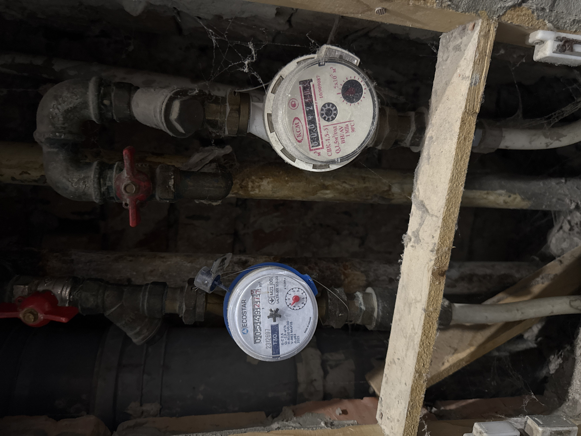 The image shows a cluttered and dimly lit space, possibly a basement or utility area, with exposed pipes and water meters. Two water meters are prominently displayed, one labeled "KCM" and the other "ECOSTAR," each connected to a network of pipes with valves, some showing signs of corrosion. Cobwebs are visible in the corners and around the pipes, adding to the unkempt appearance of the scene. A wooden support beam partially obscures the view on the right side of the frame.