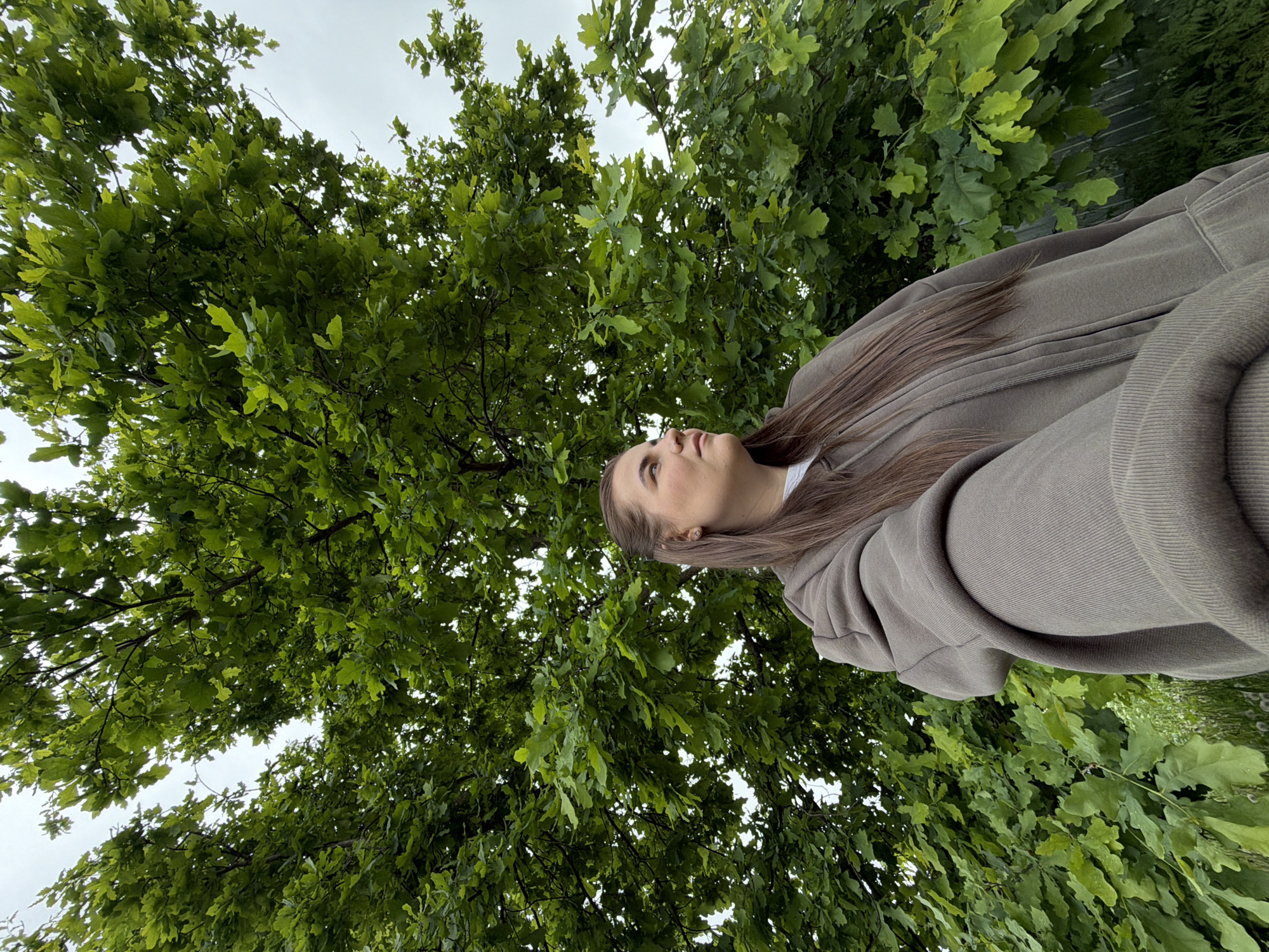 A young woman with long, straight brown hair, wearing a greyish-brown or olive-green hoodie, is captured outdoors, looking upwards. Her upper body is visible, positioned on the right side of the frame, with her head tilted to gaze skyward. The background is predominantly filled with the dense, vibrant green foliage of a large tree, likely an oak, whose lobed leaves are clearly visible. A light, overcast sky peeks through the canopy, indicating daytime with diffused light. The woman's expression is calm and contemplative as she observes the view above. The lush greenery suggests a natural setting, such as a park or forest, in Marakushi, Russia, likely during spring or summer. No text is visible in the image.