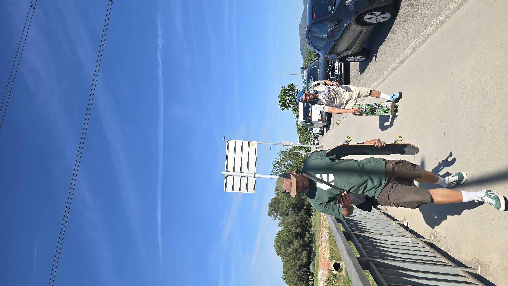 Two men with skateboards are pictured outdoors on a sunny day in Santa Perpètua de Mogoda, Spain. The image is taken from a slightly elevated perspective, looking down and along a paved path next to a road.
In the foreground, a man stands on the paved path beside a metal railing. He is wearing a dark green t-shirt with a white logo, olive-colored cargo shorts, a two-tone baseball cap, white socks, and green-and-white sneakers. He holds a black skateboard under his left arm and is looking down at a black smartphone held in his left hand. A black shoulder bag strap crosses his chest. His prominent shadow falls on the path, indicating strong sunlight.
Further along the path, closer to the road and parked cars, another man is visible. He wears a light-colored t-shirt with a graphic print, light shorts, a blue beanie, white socks, and blue sneakers. He holds a skateboard with a green graphic design under his arm and is leaning against a parked black car. He appears to be looking towards the camera, wearing sunglasses.
The setting includes a road with several parked cars, including a black car whose license plate "C 4973 MC" is partially visible. Between the two men stands a white directional sign with multiple horizontal panels. The background features lush green trees and distant hills or mountains under a bright blue sky, with visible wispy clouds and airplane contrails. The clear sky and strong shadows suggest it is midday or early afternoon. The activity appears to be a casual break during a skateboarding session.