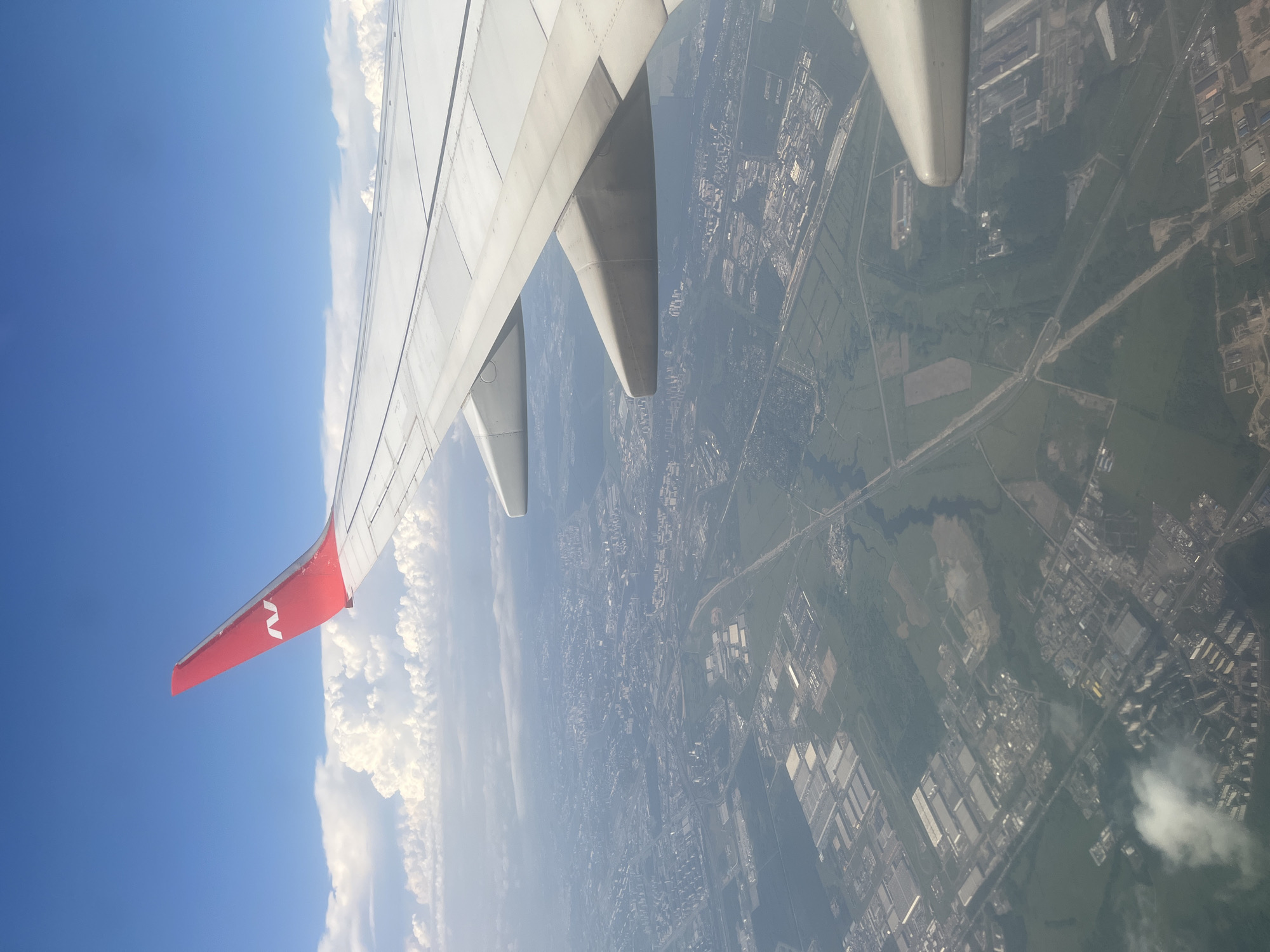 An aerial view from an airplane window shows a sprawling urban landscape with an airplane wing prominently in the foreground. The wing is white with a bright red tip featuring a white stylized 'N' logo. The airplane is flying over a city, likely Stolbishche, Russia, given the distinctive industrial and residential architecture visible. Below the wing, a dense urban fabric unfolds, characterized by numerous multi-story buildings, industrial complexes with large rectangular roofs, and green spaces. Roads and what appear to be railway lines crisscross the terrain. The sky is a vibrant blue, dotted with fluffy white cumulus clouds. The overall impression is a clear, daytime shot from high altitude, capturing the transition from industrial outskirts to residential areas within a city.