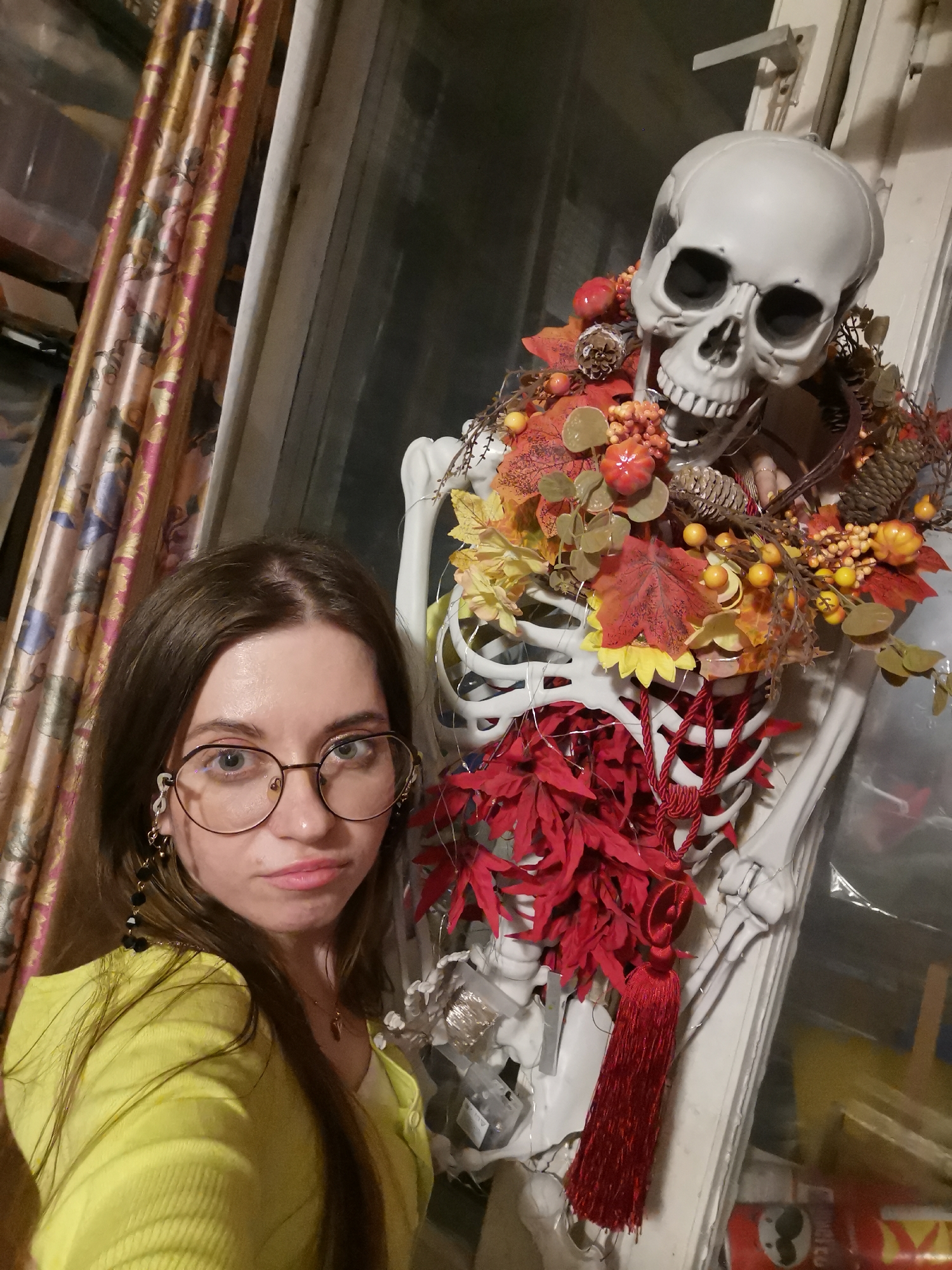 A woman poses for a selfie next to a skeleton decorated for Halloween or a similar autumn-themed event. The woman, fair-skinned with shoulder-length brown hair and wearing large, round spectacles, is positioned on the left side of the frame. She is wearing a yellow cardigan over a white top and has dangling earrings, and a necklace is visible.
To the right of the woman is a full-sized plastic skeleton adorned with an autumn-themed wreath around its neck and chest. The wreath includes artificial leaves, berries, miniature pumpkins, and pine cones. A red tassel-like decoration hangs from the skeleton's chest. The skeleton is positioned against a window frame.
The background features a patterned curtain on the left and a cluttered area with various items, some in clear plastic packaging, visible behind the skeleton.
Given the level of detail and the cluttered nature of the background, the setting appears to be indoors, possibly in a residential area. There is no information within the image to link the image to the city of Moscow, Russia.