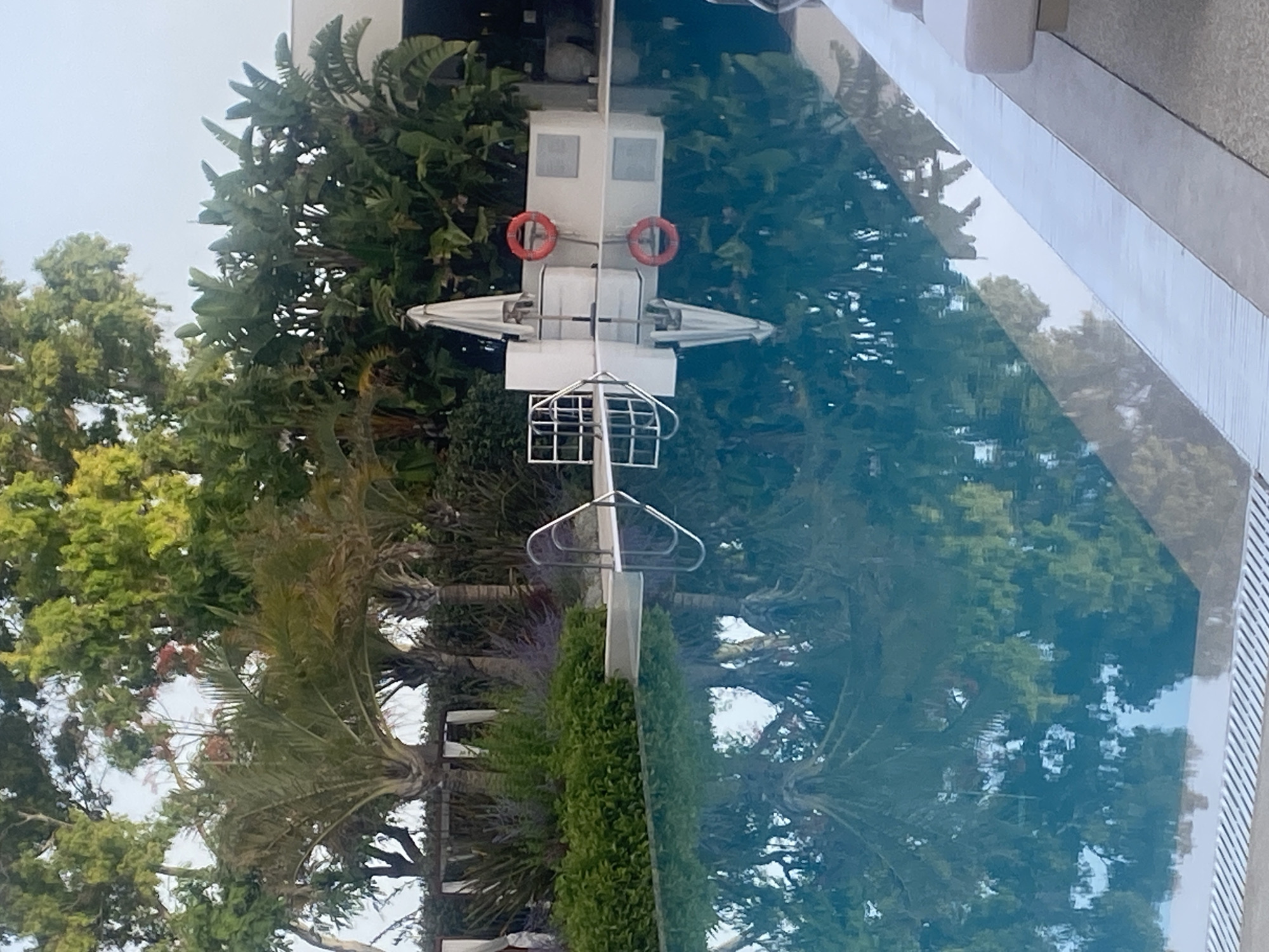 The image displays a swimming pool with clear blue water. The pool is surrounded by lush green vegetation, including palm trees and other tropical plants, which are reflected in the still water. On the right side of the pool, there is a white structure with a closed white umbrella and a red life preserver hanging on it. A metal ladder extends into the water from the left side of the pool. The pool area appears to be an outdoor space, possibly a resort or a private residence, with lounge chairs partially visible at the edge of the frame on the right. The overall scene suggests a tranquil and luxurious environment. The location specified, Resinera-voladilla, Spain, is a small village in the province of Valladolid. However, there are no specific visual cues in the image that definitively place it in this exact location.