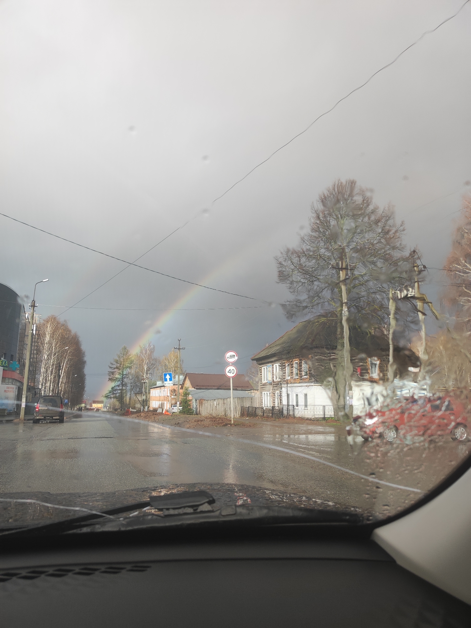 The image, taken from inside a vehicle, shows a street scene in Votkinsk, Russia on a rainy day with a rainbow visible in the distance. The road is wet and reflects the surroundings. A gray car is traveling in the same direction as the photographer's vehicle. On the right side of the road is a traditional Russian wooden house with a dark roof, and in front of it is a 40-speed limit sign and a directional sign. Power lines stretch across the sky. Rain droplets are visible on the windshield of the vehicle.