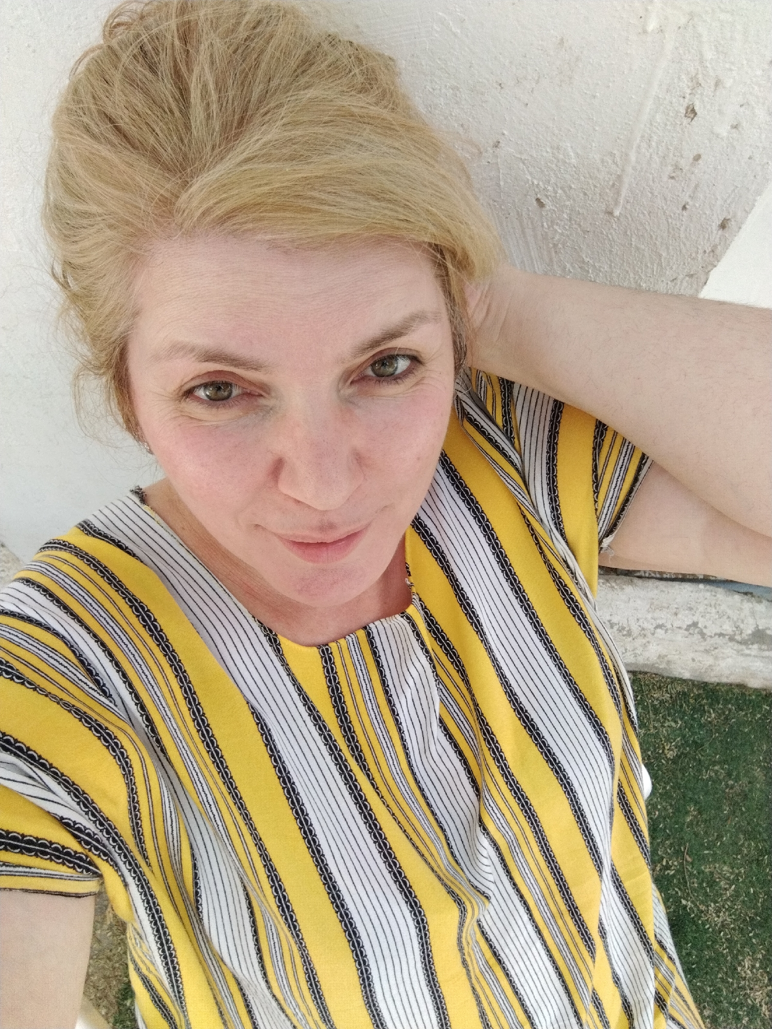 A woman is shown in an eye-level, close-up portrait in Al Mas'oudi, United Arab Emirates. She has fair skin, green eyes, and blonde hair piled on her head. She wears a yellow and white striped shirt. Her right arm is raised, hand on the back of her head. She stands in front of a white concrete wall with green grass in the lower right corner.