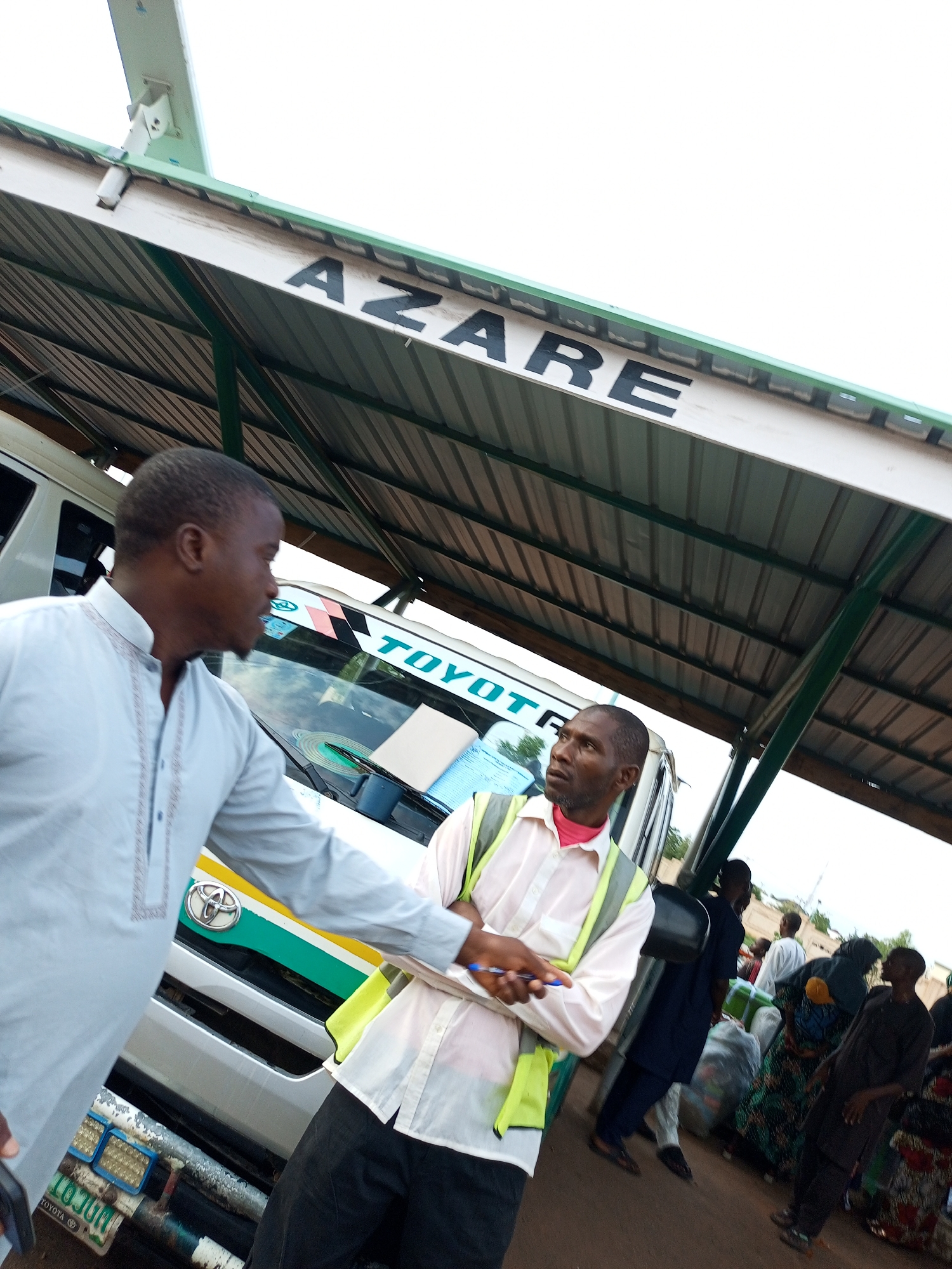 The image depicts a busy bus park under a covered shelter in Azare, Gombe State, Nigeria. Two men are the primary subjects, engaged in conversation. The man on the left, wearing a light blue traditional garment, stands near the front of a white Toyota bus. The man on the right, dressed in a pink shirt and a high-visibility green vest over dark pants, faces the man on the left. The bus has "TOYOTA" prominently displayed on its windshield. In the background, several other people are visible, some carrying luggage, suggesting active travel. The environment is bright, likely during daytime, with the overcast sky visible above the shelter. The shelter itself is constructed with a corrugated metal roof supported by green metal posts. License plates are visible on the bus.