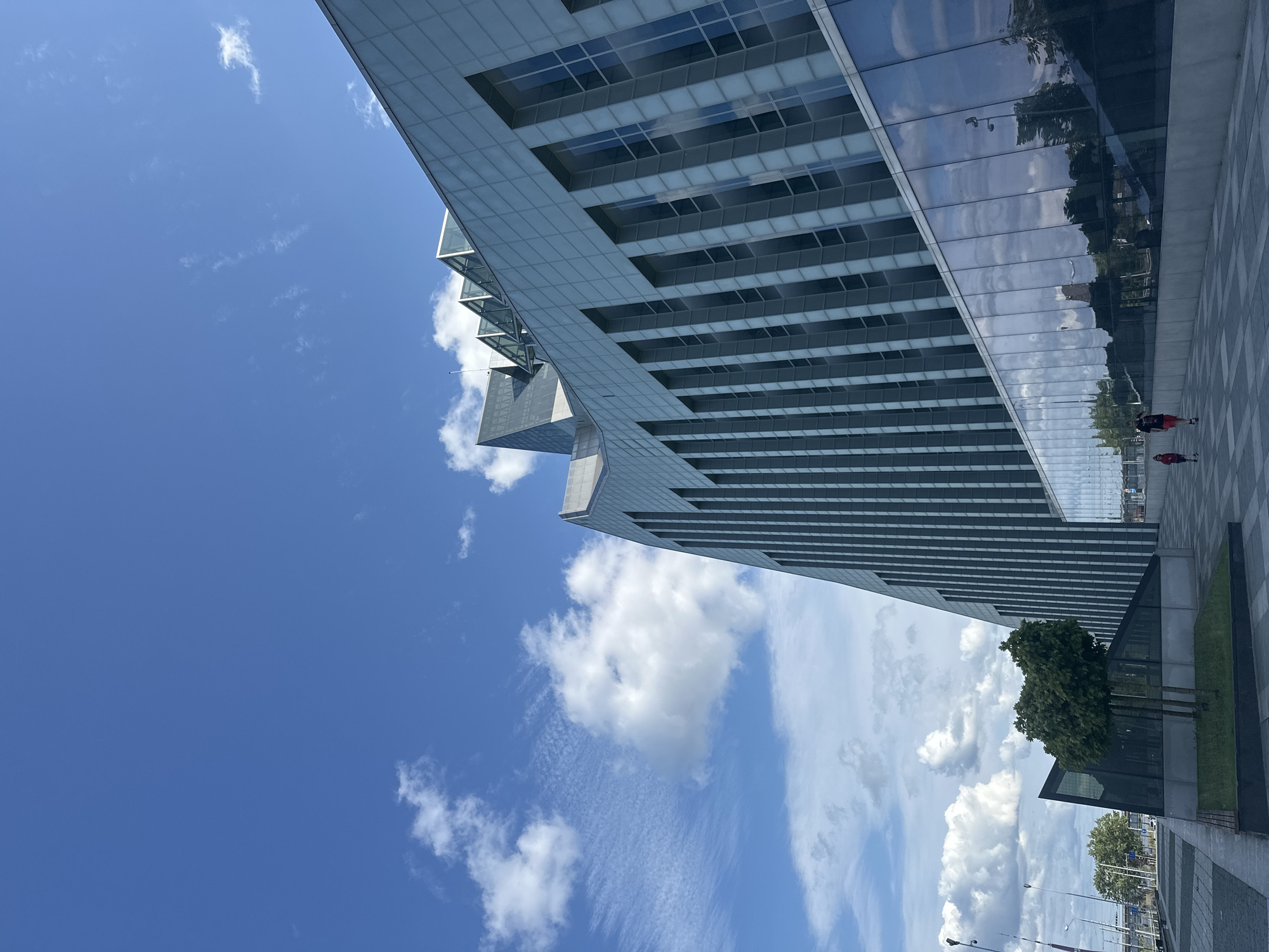 The image depicts a modern architectural scene in Riga, Latvia, under a bright blue sky with scattered white clouds. The primary subject is a striking, multi-faceted glass and concrete building with a distinctive angled design. The building's facade reflects the sky and surrounding environment, creating a shimmering effect.
In the foreground, a paved walkway with a checkered pattern leads alongside the building. Two individuals, seen from a distance, are walking on the pavement, one wearing a red jacket. A small, manicured tree adds a touch of greenery to the scene. Further in the background, more trees and streetlights are visible, suggesting an urban environment.
The perspective is from a low angle, looking up towards the expansive sky and the imposing structure, emphasizing its scale and modern design. The time of day appears to be daytime, likely late morning or early afternoon, given the clear, bright sunlight. The weather is fair and sunny, with a mix of fluffy and wispy clouds. There is no visible text in the image. The overall impression is one of contemporary urban design and pleasant weather conditions.
