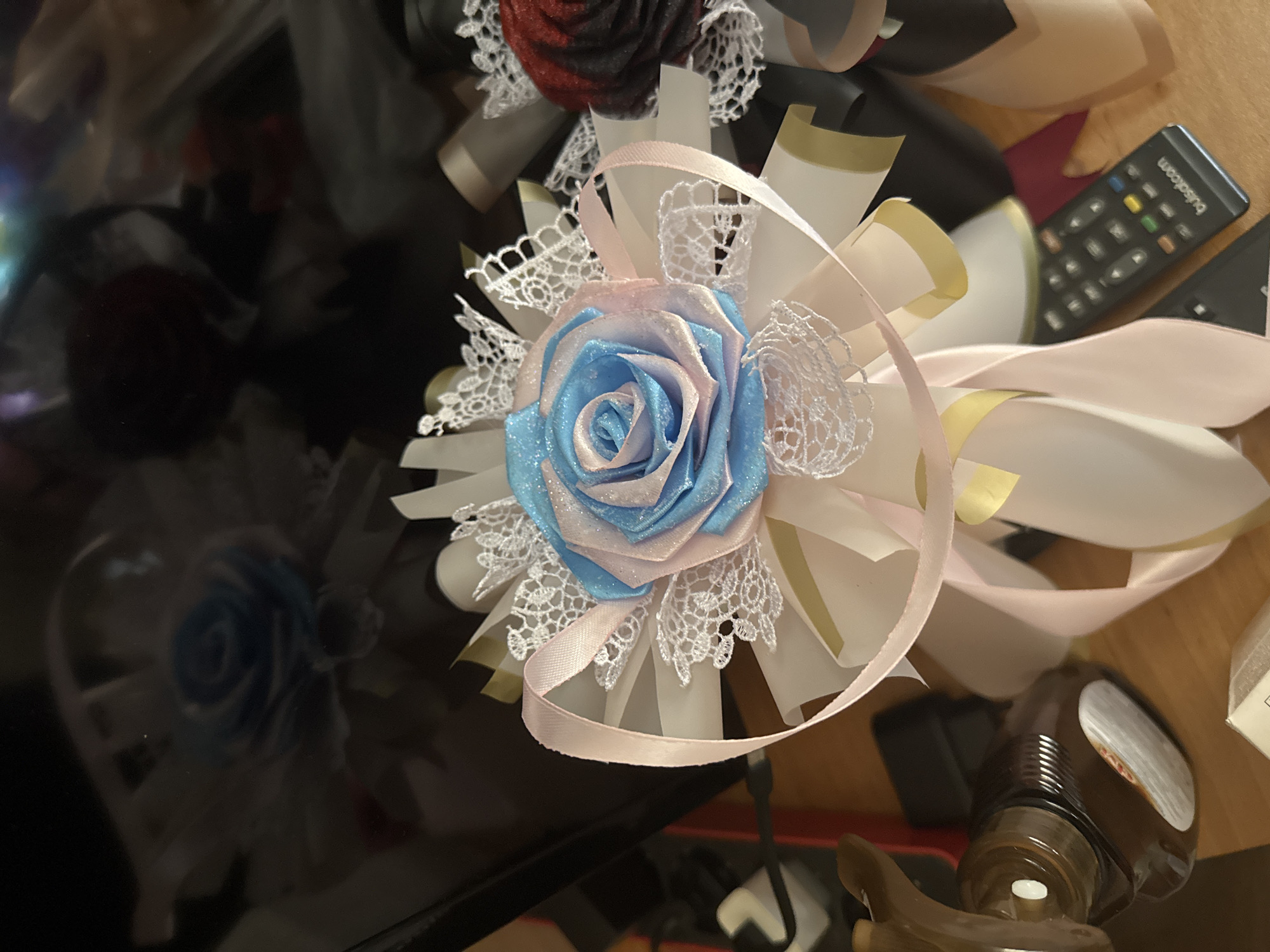 This is a close-up, eye-level shot of decorative fabric roses, possibly part of a bouquet or arrangement, placed on a wooden surface. The main subject is a rose crafted from shimmering blue and pink fabric, with white lace trim and light pink ribbons encircling it. Behind this rose, another rose, dark red in color, is partially visible. The background is dark and reflective, showing indistinct shapes that appear to be more of the same decorative elements. A brown glass bottle with a white cap and a label is positioned to the left of the arrangement. To the right, a black remote control with colorful buttons rests on the surface. The overall lighting is dim, with a slight glare on the reflective background. There is no indication of the city of Sredets, Bulgaria.