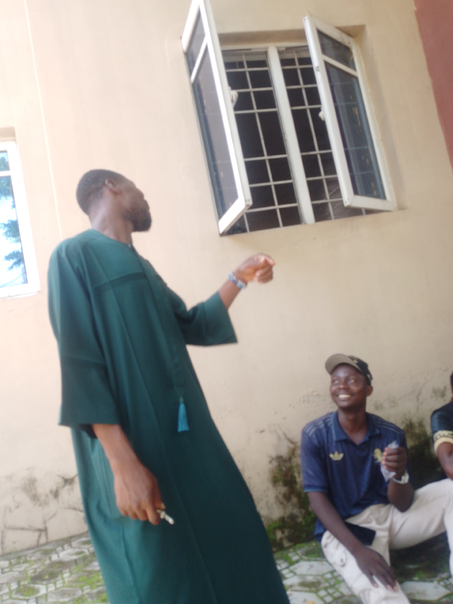 This media file captures an outdoor scene with at least three individuals. The primary subject, in the foreground, is a man wearing a long, dark green robe with wide sleeves and a decorative blue tassel. He is facing away from the camera and appears to be gesturing with his right hand, which is adorned with a blue beaded bracelet. He is also holding a small, silver object in his hand.
To the right of the main subject, another person is seated and looking upwards with a broad smile, suggesting happiness or amusement. This individual is wearing a dark blue polo shirt with a visible logo, a baseball cap, and light-colored pants. They are holding a white object in their hand. A third person is partially visible on the far right, wearing a dark shirt with a gold and black pattern.
The setting appears to be outdoors, possibly a courtyard or a public space, with a beige-colored building in the background. A window with white frames and a grid pattern is visible on the building, with one section open. The ground is paved with interlocking bricks, and some patches of moss or greenery are visible. The lighting suggests it is daytime, with natural light illuminating the scene. The overall mood seems relaxed and conversational. No visible text is present in the image.