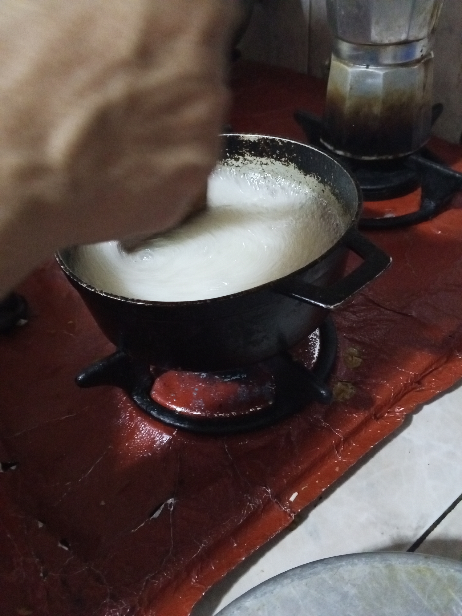 The image shows a person stirring a white liquid in a black pot on a gas burner. Another gas burner with a moka pot is visible in the background. The cooking area is covered by a red sheet or cloth. The floor appears to be tiled, with some tiles visible in the lower right corner. The location is indicated as Tinaquillo, Venezuela.