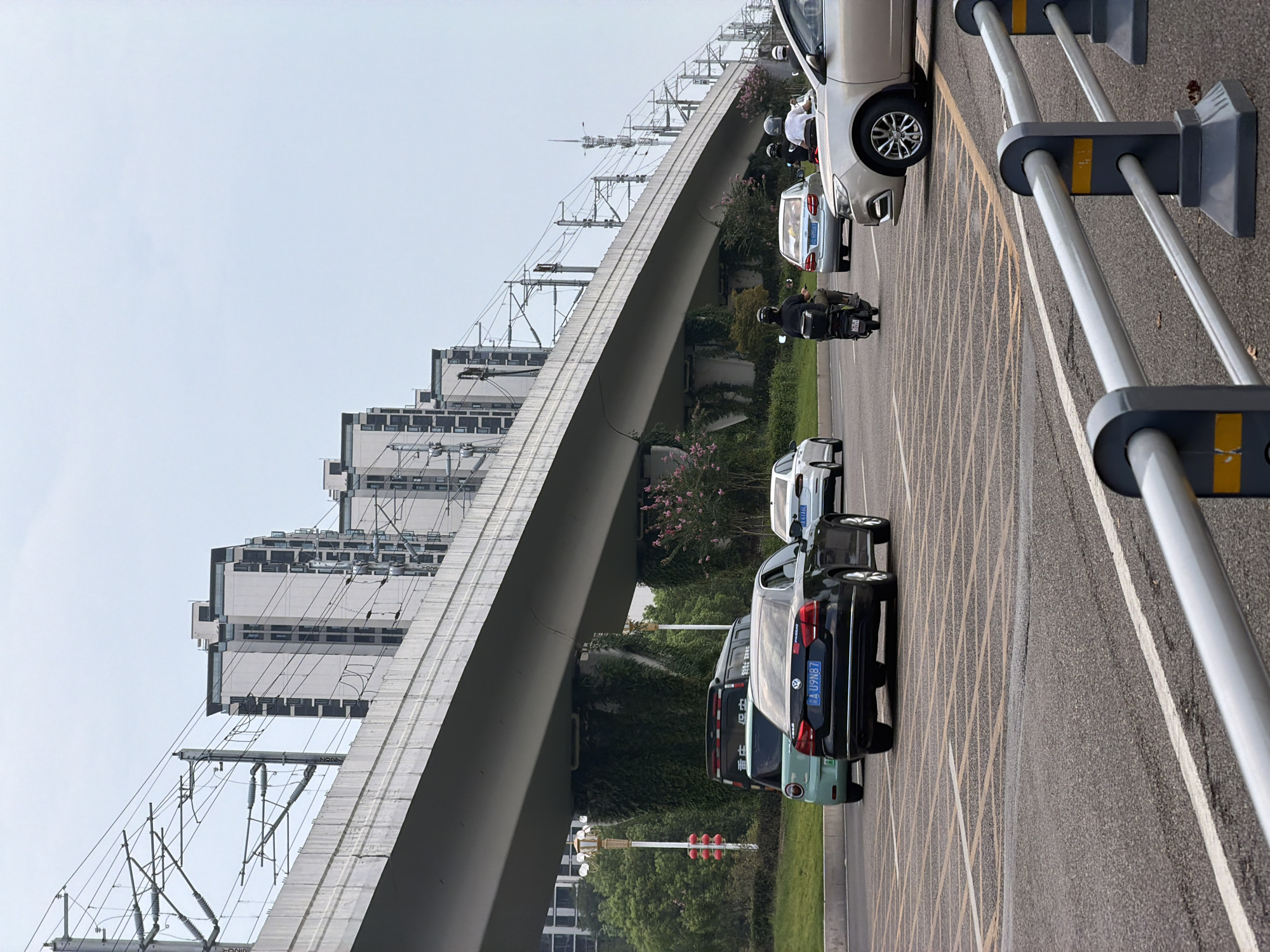 In Jiangjin, China, a street scene unfolds beneath a massive elevated transportation structure, likely a highway or railway. The structure casts a long shadow over the roadway below. On the road, several vehicles are in motion. A black sedan with a blue license plate reading "A U9N87" is prominent in the foreground, heading away from the viewer. To its left, a small, light green car is visible. Further down the road, a silver sedan and a scooter with a rider wearing a helmet are also moving. A white car is seen in the distance. The environment includes green foliage on the supporting pillars of the elevated structure and trees lining the side of the road, some with pink blossoms. The sky is overcast. In the background, several modern, multi-story buildings with dark window sections rise up, interspersed with power lines and poles that run along the elevated structure. The scene appears to be a typical urban setting with active transportation infrastructure and vehicular traffic.