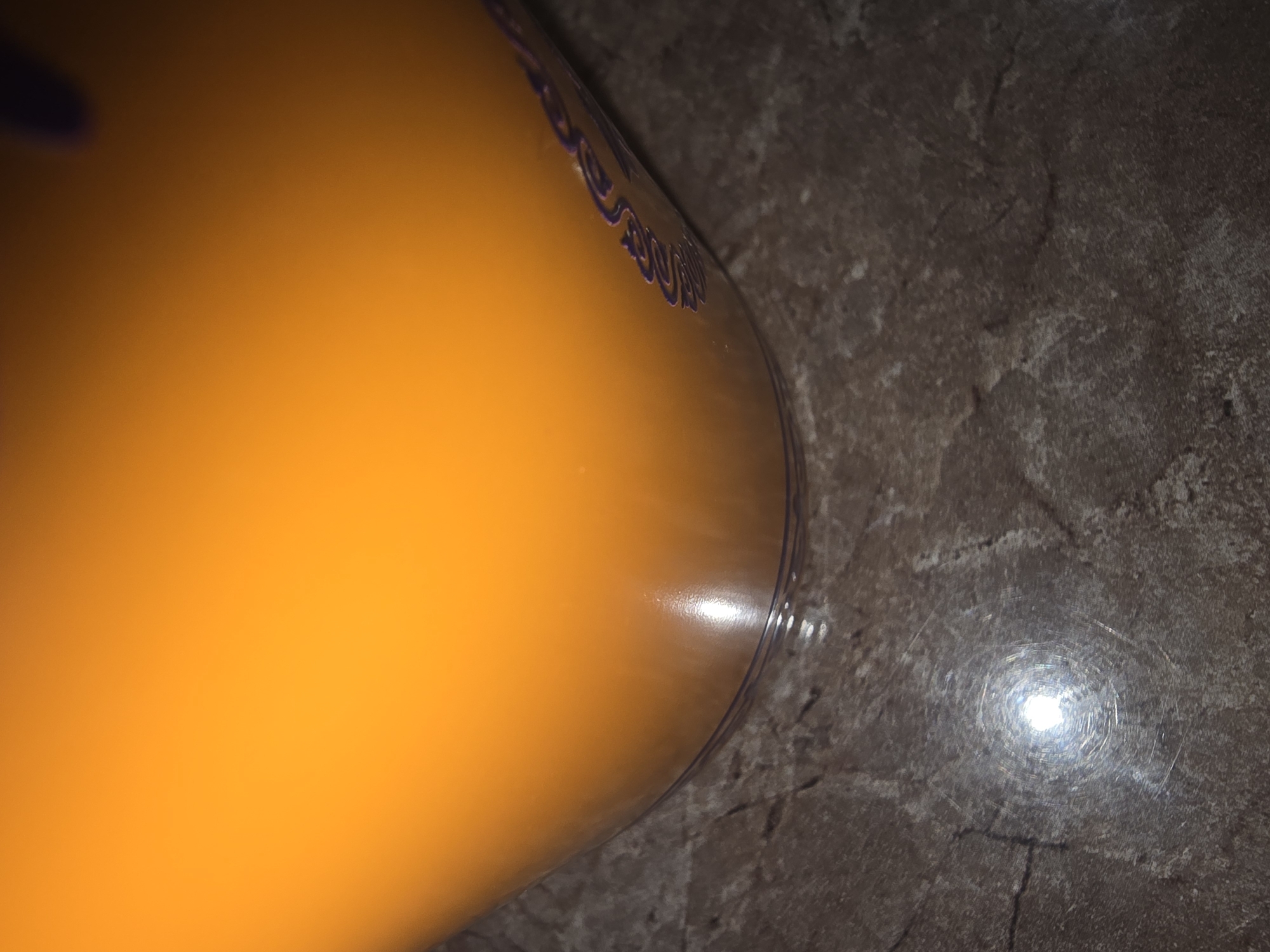 The image shows a close-up of a transparent container filled with an orange liquid. The container has a textured pattern or branding in purple along its side. The surface the container is resting on appears to be a dark, mottled countertop or table. A bright, circular reflection is visible on this surface, possibly from a light source. The scene is indoors, and the overall lighting is dim, with the bright orange liquid being the most illuminated element. There is no discernible indication of the location being in the city of Ulan-Ude, Russia.