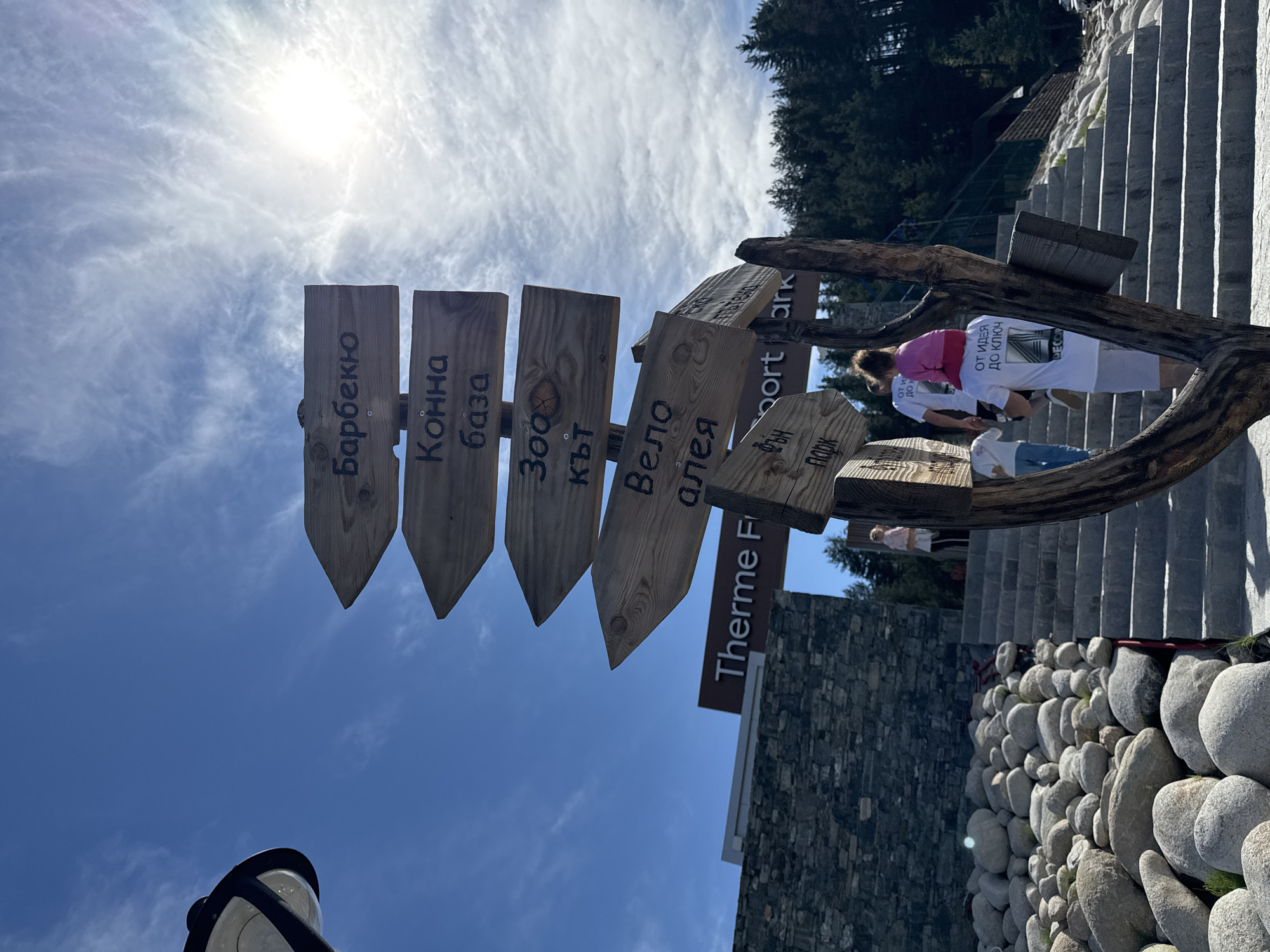The image shows a wooden signpost pointing in multiple directions, possibly in a recreational or resort area. The signs, with their pointed ends, have text written in a Cyrillic alphabet, likely Bulgarian. The first sign says "Барбекю," the second says "Конна база," the third says "3000 кът," and the fourth sign says "Вело алея." One of the signs says "Sport Park."
A partial sign is visible at the bottom of the frame with the words "Therme F." A group of people is walking up a set of stairs, one of them carries another one on their shoulders.
The background includes a stone wall, trees, and a bright, partly cloudy sky. Below the staircase, there is a wall of round, grey rocks. A lamp post is visible at the bottom left corner of the frame. The location is somewhere in a park or resort, possibly near a thermal spa.