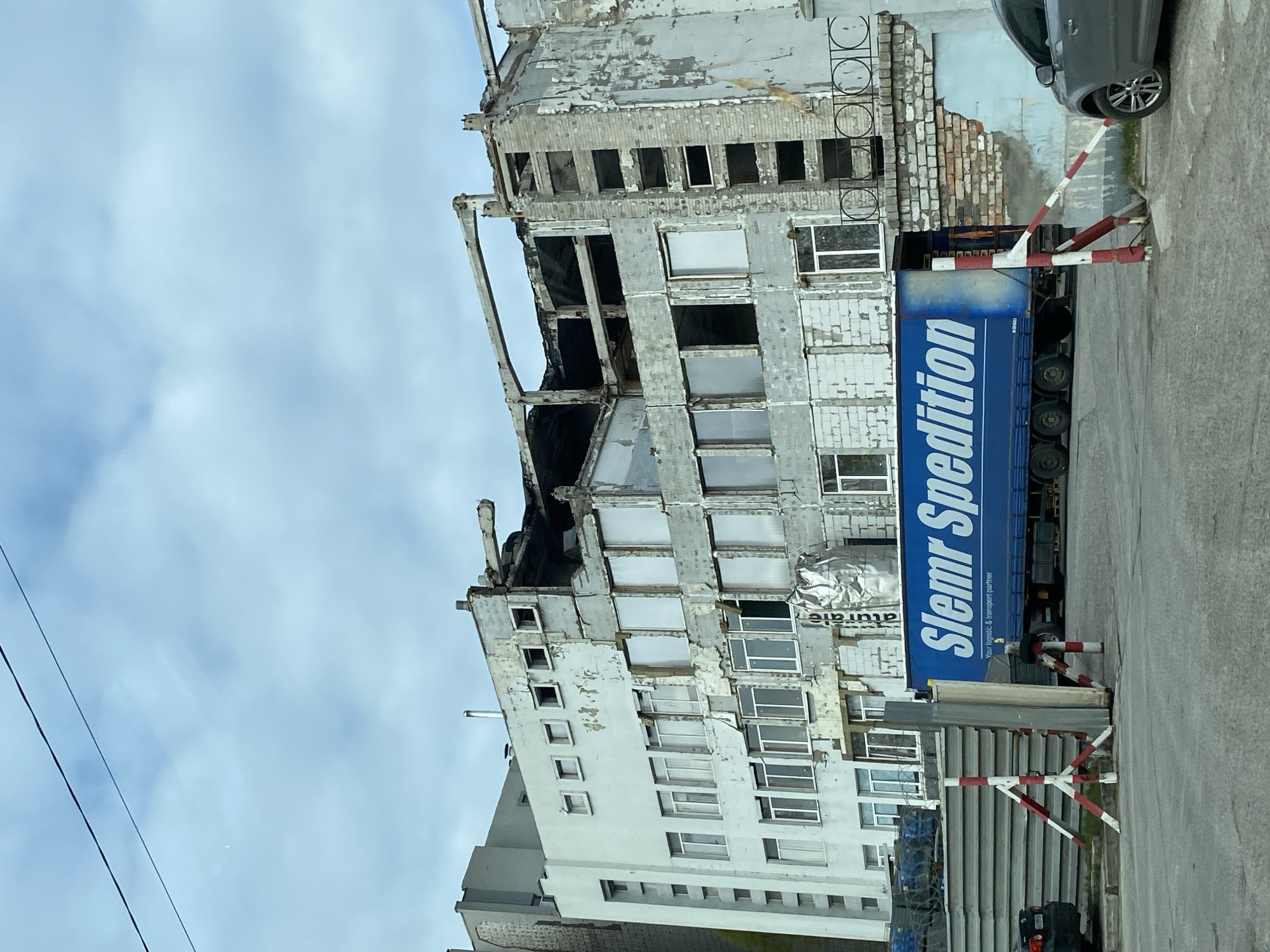 The image depicts a partially demolished building in an urban environment. The building's facade is heavily damaged, with exposed brick, missing windows, and peeling paint. A large blue truck with "Slemr Spedition" written on its side is parked in front of the building. There are red and white striped barriers nearby. A gray car is visible in the background. The sky is overcast with a mix of white and blue clouds. Overhead power lines are visible in the top left of the image. The surrounding environment appears to be a mix of concrete and pavement.