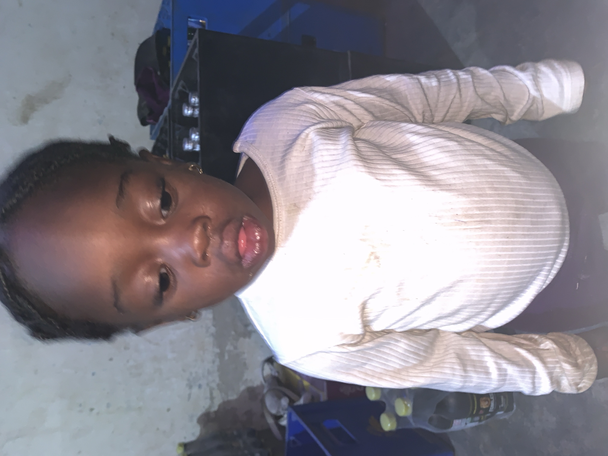 A child with dark skin and braided hair wears a long-sleeved white ribbed shirt. The child is indoors, as suggested by the background which includes a mottled white wall and a partially visible dark cabinet or shelving unit. Items such as bottles and a blue crate are present on the floor near the bottom edge of the frame. The photo appears to be taken from a slightly elevated angle. It's impossible to confirm the location in the city of Kabong, Nigeria based only on visual information.