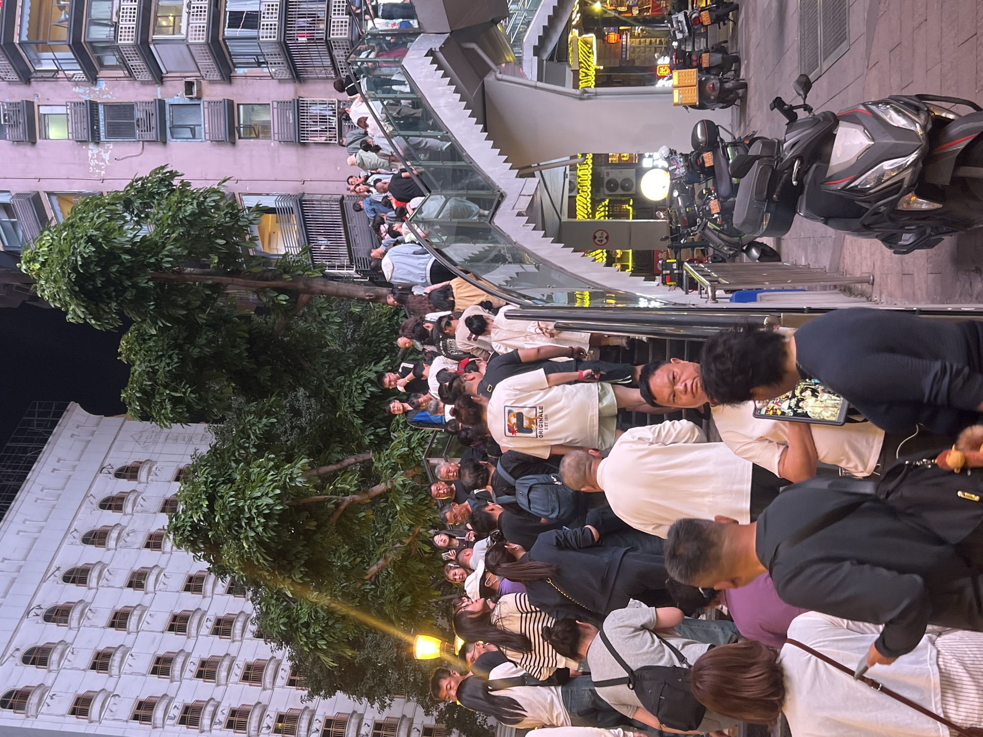 A crowd of people stands on a narrow sidewalk next to a building and stairs leading to a higher level. Many are looking in the same direction. The building on the left is light-colored with many windows. There are also buildings in the background on the right. Several parked motorcycles are along the edge of the sidewalk on the right. Tall trees are visible between the buildings. This is located in Yuzhong District, China.