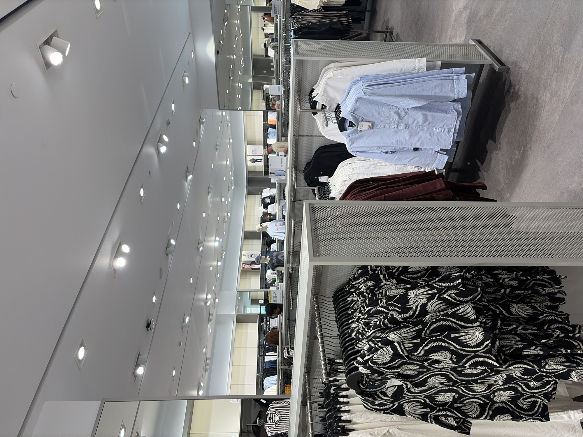 The image depicts the interior of a retail clothing store, likely a department store or a fashion chain, in Kuta, Indonesia. The store features a brightly lit environment with numerous ceiling lights. Racks of men's clothing are prominently displayed, including shirts of various colors and patterns, such as striped, floral, and solid blue. Some clothing items are on hangers, while others are folded or draped. In the background, mannequins are visible, showcasing outfits. A few individuals, presumed to be customers or staff, are present in the store, with one person appearing to be browsing. The store's flooring is a muted gray, and the overall aesthetic suggests a modern and organized retail space.