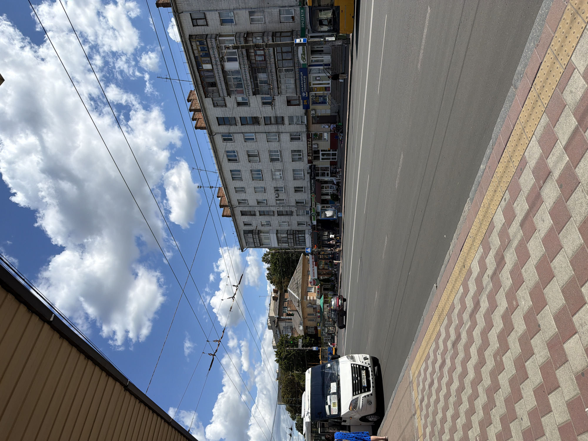 A wide-angle, eye-level shot captures a street scene in Zhytomyr, Ukraine. A white minivan with registration number AM 2866 CH is parked at the curb on the left, facing right. A woman in a blue dress stands near the front of the minivan. A red car is in the middle lane of the multi-lane road, moving away from the viewer. On the right side of the street, a yellow bus is visible. The buildings lining the street are multi-story, with the closest being a light-colored apartment building with numerous windows and balconies. Shops are visible at the ground level of the apartment building, with signs in Cyrillic script. Overhead, power lines crisscross the sky, which is blue with scattered white clouds. The sidewalk in the foreground is paved with alternating red and beige rectangular tiles, with a yellow tactile paving strip. The road is asphalt.