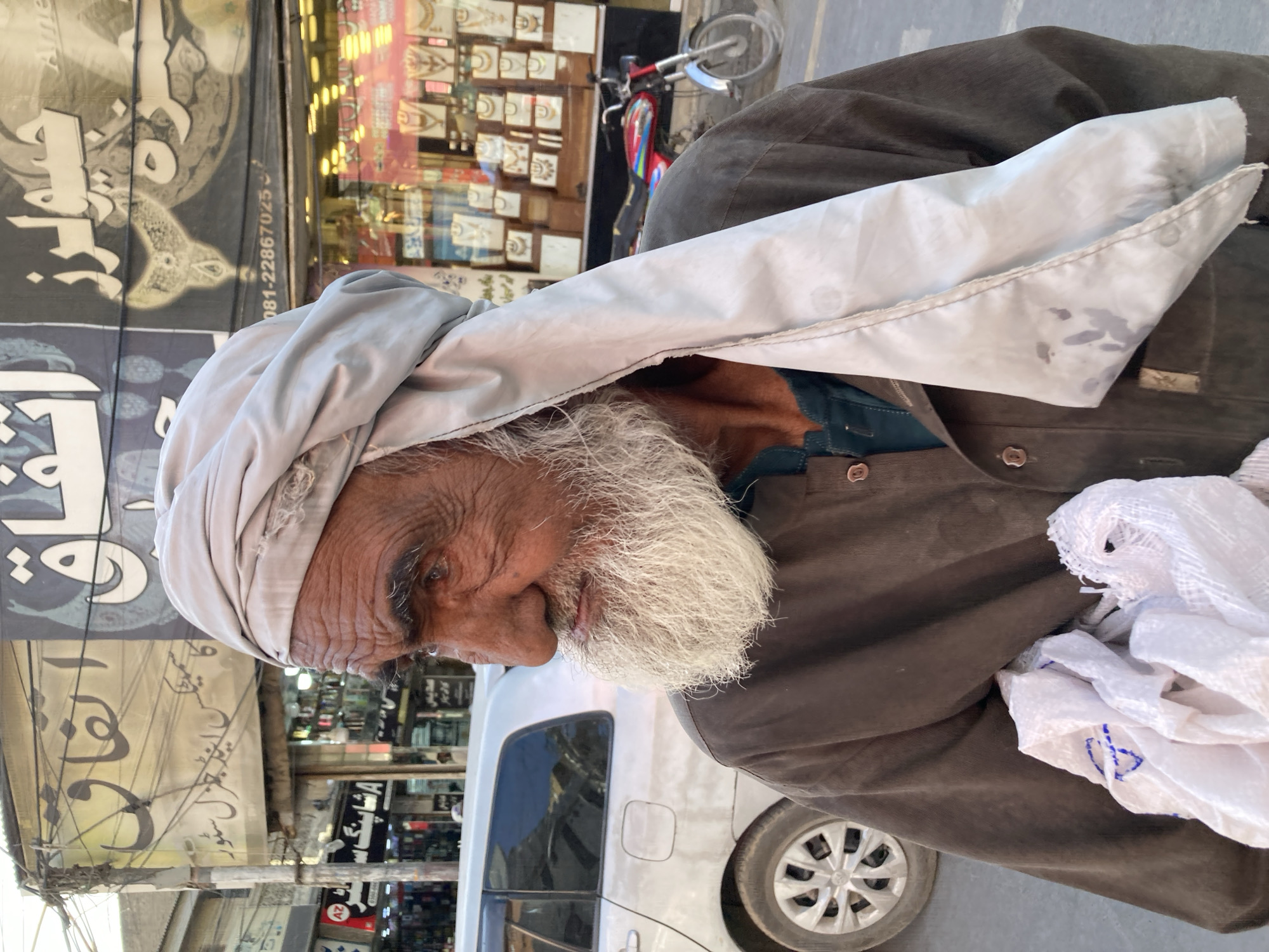 An older man with a white beard and head covering is the main subject of this outdoor, portrait-style shot. The man is wearing a brown jacket and holding a white cloth or bag. The location appears to be a city street, evidenced by the storefronts, cars, and other urban elements visible in the background. The storefronts display signs and merchandise. A motorcycle is partially visible, parked near one of the stores. A white car is parked next to the man. The image was captured in Quetta City Tehsil, Pakistan, as indicated by the signs.