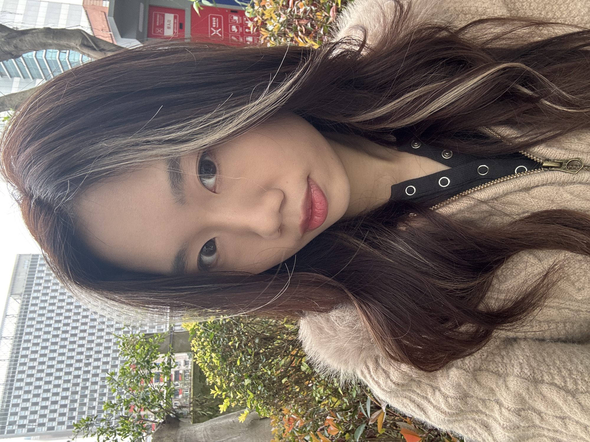 A single East Asian female subject is centered, occupying the majority of the frame, and looking directly into the camera. She has long, wavy dark brown hair with prominent light blonde streaks framing the right side of her face (viewer's left). Her eyes are dark, her eyebrows defined, and her lips are colored with a reddish-pink hue and are slightly parted. She wears a textured, light beige cable-knit outer garment, possibly a sweater or cardigan, with a prominent furry or fuzzy collar. Beneath this, a dark-colored top with silver-toned snap buttons is visible, partially fastened by a brass-colored zipper. The outdoor background features dense green and russet-toned foliage. To the left, a portion of a tree trunk is visible. To the right, a red public utility kiosk with white Chinese characters and red 'X' symbols is partially visible. In the distant background, urban architecture includes a multi-story building characterized by a grid of uniform windows, and a taller structure with blue-tinted glass panels. The scene is situated in Nan'an District, China.