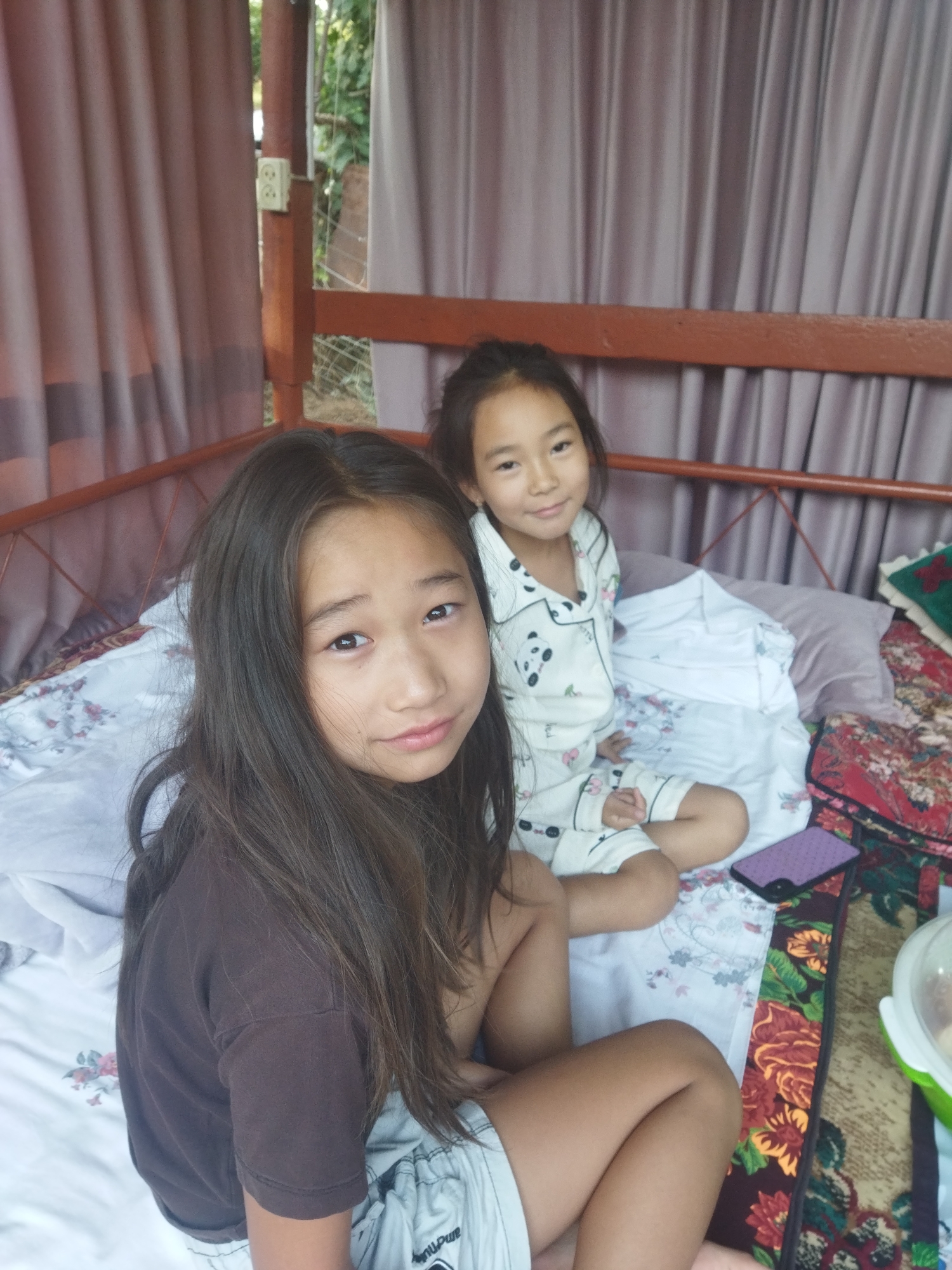 Two young girls are seated on a bed. The girl in the foreground has long, dark brown hair and is wearing a brown t-shirt and light-colored shorts. She is looking towards the camera with a neutral expression. The girl in the background has dark hair pulled back and is wearing pajamas with panda and cherry patterns. She is looking slightly off-camera and smiling. The bed is covered with patterned sheets and pillows. A purple phone is visible on the bed to the right of the girls. Behind them, a curtain is drawn, and a wooden structure is visible. The setting appears to be outdoors, possibly on a porch or balcony, with greenery visible in the background, suggesting a location in the city of Mailuu-Suu City, Kyrgyzstan.