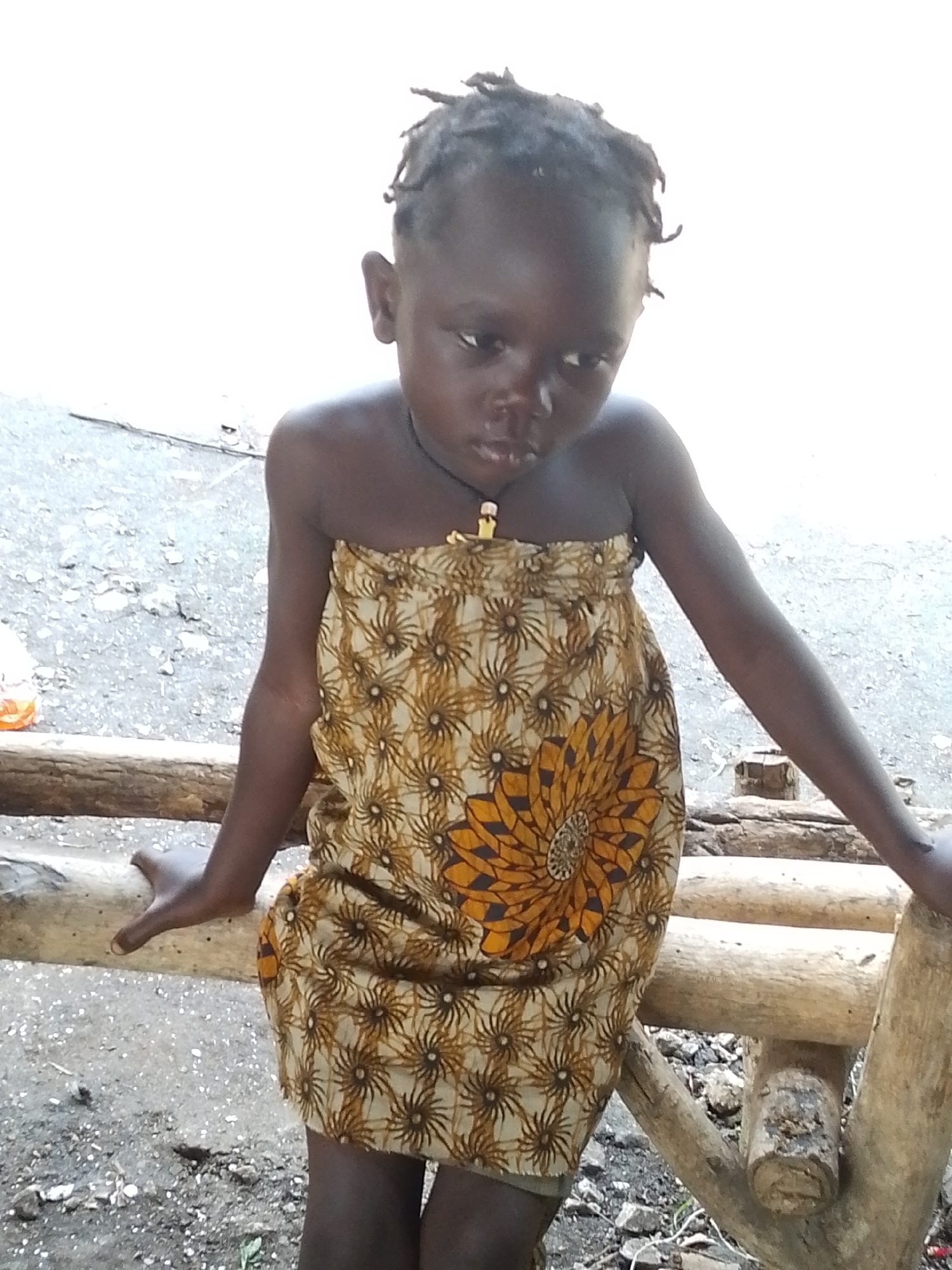 A young Black child, with dark skin and dreadlocks, is seated on a wooden structure, possibly a bench or fence, in an outdoor setting. The child is wearing a strapless, tan and brown patterned dress with a large floral design on the front. A dark necklace with a yellow pendant is visible around the child's neck. The child is looking downwards and to the right. The background consists of a mottled gray surface, possibly dirt or gravel, with scattered small rocks and debris. The lighting is bright, suggesting daylight. The overall scene appears to be in a rural or semi-rural area, possibly in Otulu, Nigeria, given the context.
