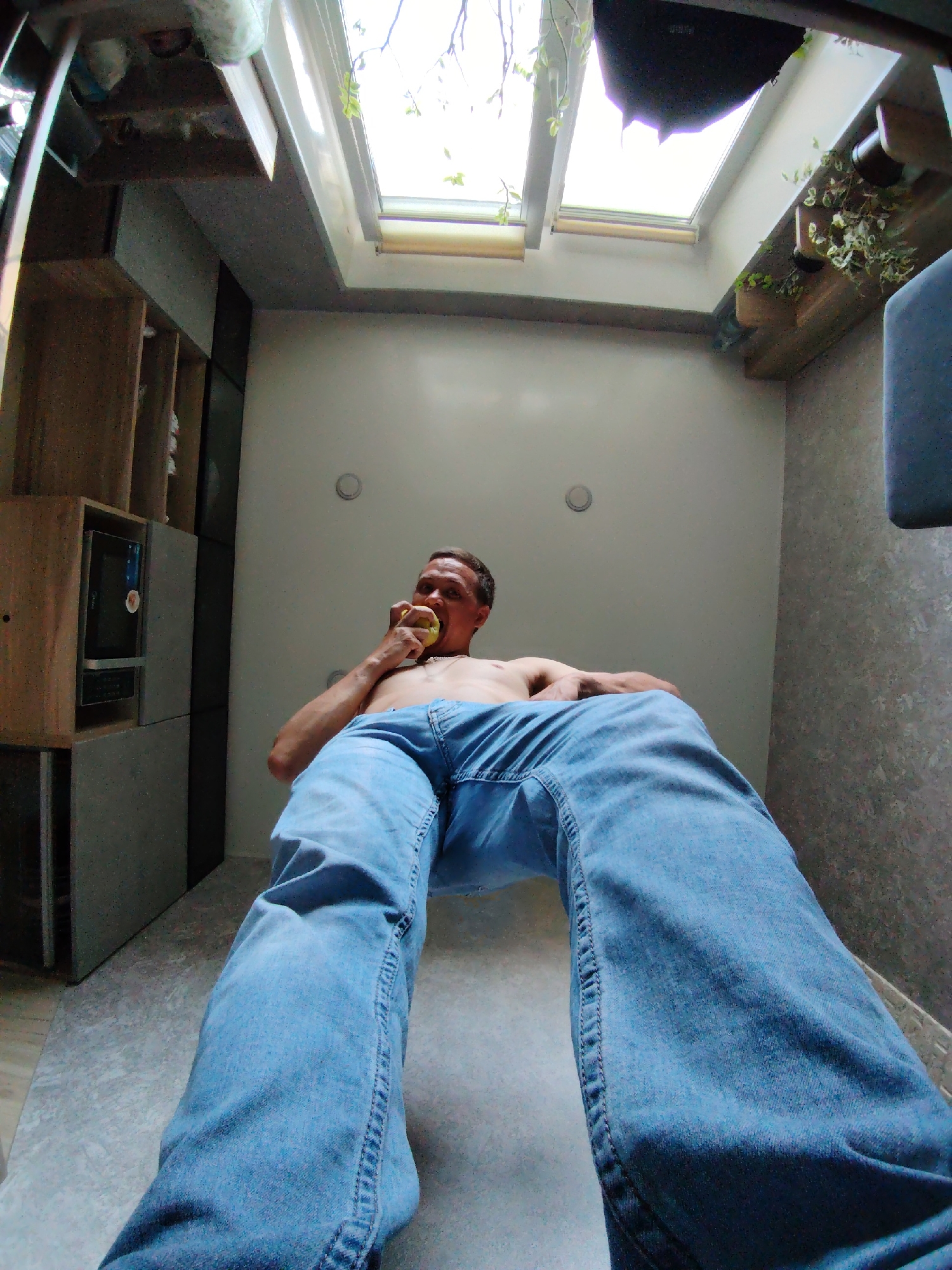 The image shows a man wearing blue jeans, sitting indoors and eating an apple. He is shirtless. The room appears to be a modern kitchen or living area with a skylight and built-in cabinetry. The camera angle is low, looking up at the man and his surroundings. There is no visible indication of the city of Chaikovsky, Russia.