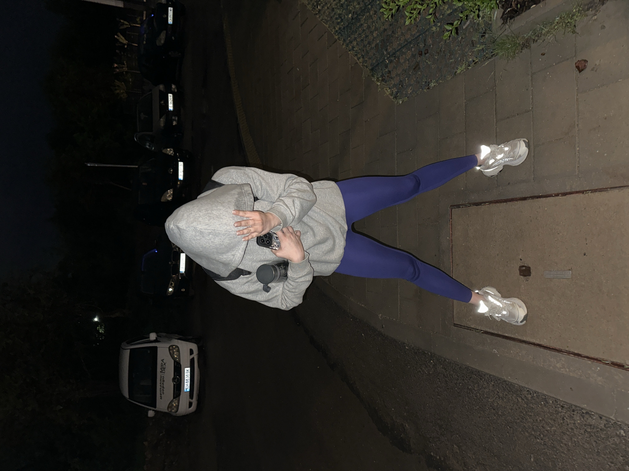 A photograph shows a person wearing a grey hoodie and blue leggings standing on a sidewalk. The person's face is obscured by the hoodie, and they are holding a phone and a water bottle. Their shoes are reflective sneakers. The sidewalk is made of concrete slabs. To the left, there is a street with parked cars and a white van with "ВЕГА" written on its side in Cyrillic, indicating the location is Sofia, Bulgaria. The lighting suggests it is nighttime or dusk. Vegetation is visible along the edge of the sidewalk.