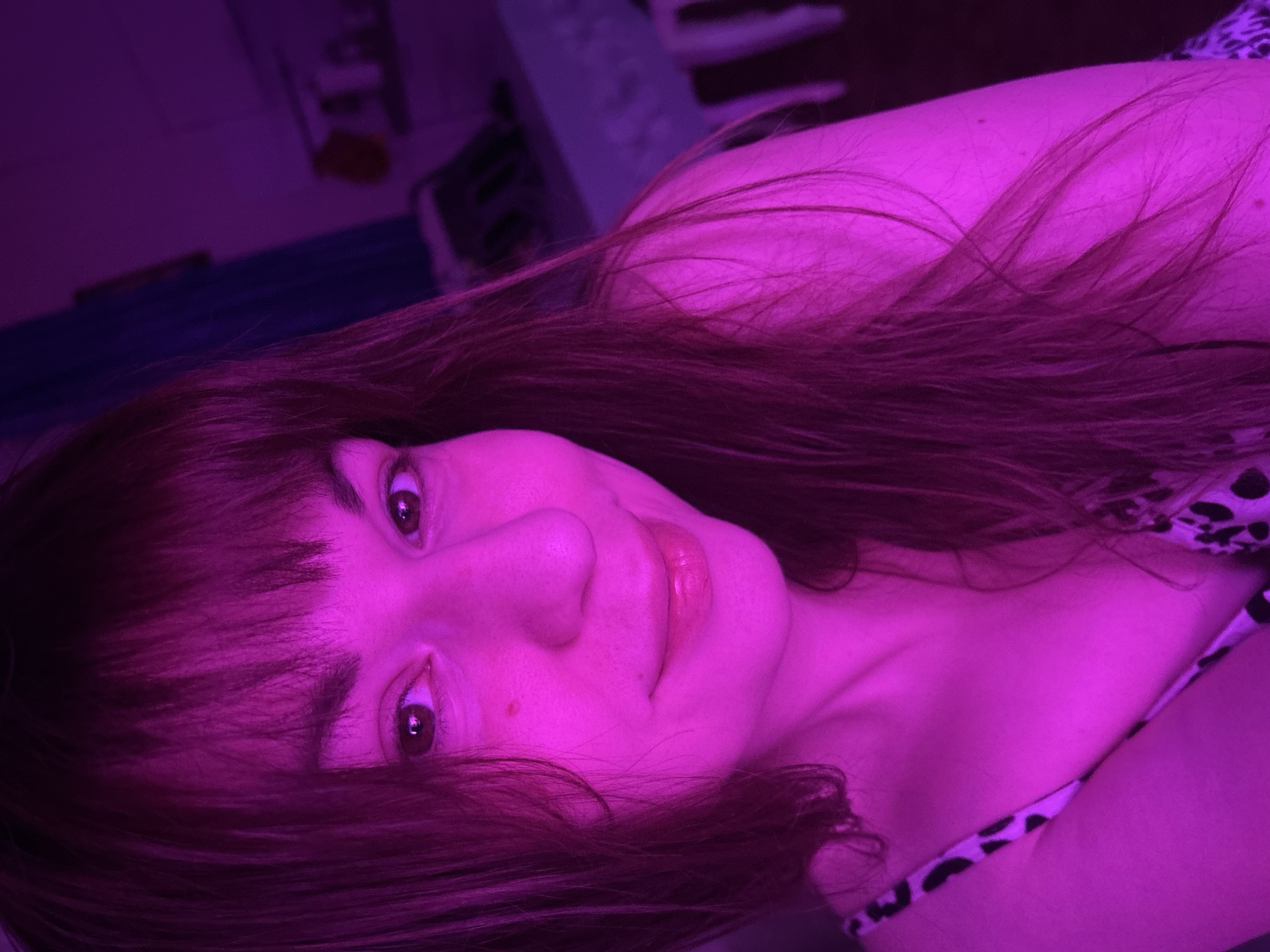 This image is a portrait of an adult female, illuminated by a dominant magenta-pink light source, appearing to be an indoor photograph. The subject occupies the majority of the frame, facing the viewer with a slight head tilt and a subtle smile. Her hair is dark, styled with bangs across her forehead, and long strands fall over her shoulders and chest. Her lips appear glossy. A small mark is visible on her right cheek. She is wearing a garment with a black and white animal print, visible on her left shoulder and upper chest. The background is out of focus and heavily saturated with the same magenta-pink light, revealing indistinct shapes consistent with typical room furnishings, possibly including a wall or curtain segment behind her left side. The specific city of Фоки, Russia, cannot be confirmed from the visual evidence provided in the image.