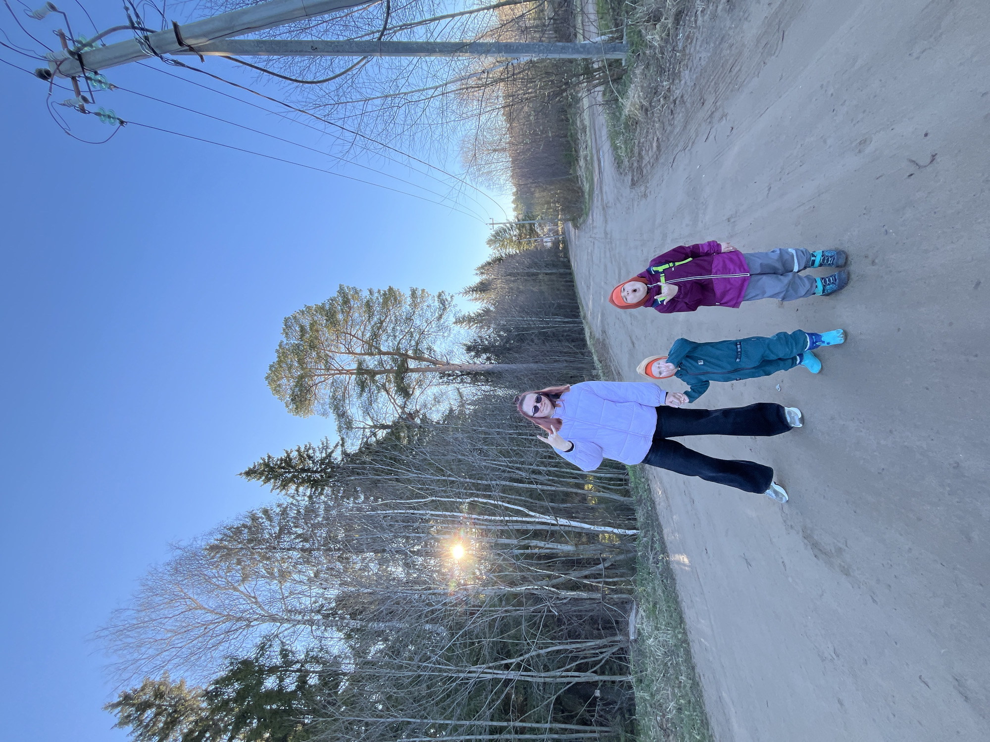 The image shows three people on a dirt road, possibly in a rural or semi-rural setting in Chaikovsky, Russia. Two children wearing winter clothes and hats are lying on the road. An adult woman in a light purple jacket and dark pants stands near them, holding up two fingers in a peace sign. The background features trees, a clear blue sky, and utility poles with wires. The sun is visible through the trees, suggesting it might be late afternoon or early morning.