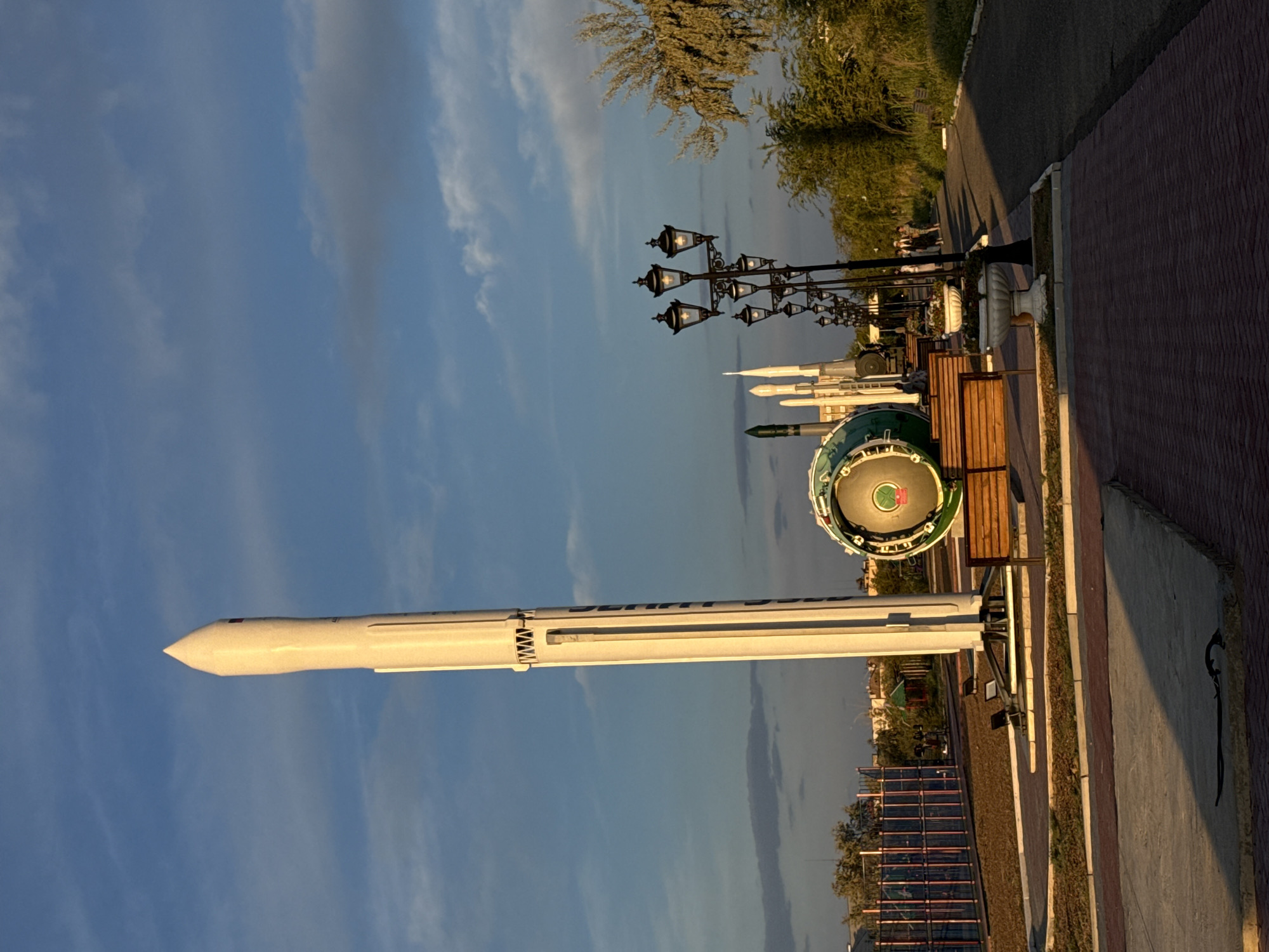 The image depicts a clear sky with scattered clouds over an outdoor display. Dominating the scene is a tall, white rocket model standing vertically. To its left, part of a circular, green and metal object, possibly another rocket component or exhibit, is visible. In the background, more rocket-like structures and what appears to be a playground with colorful fencing can be seen. A pathway lined with decorative lampposts runs through the scene, with a few people walking along it. The area is landscaped with trees and bushes, and benches are placed near the exhibits. The overall impression is of a public space dedicated to space exploration, likely located in Baikonur, Kazakhstan, given the presence of rocket exhibits.