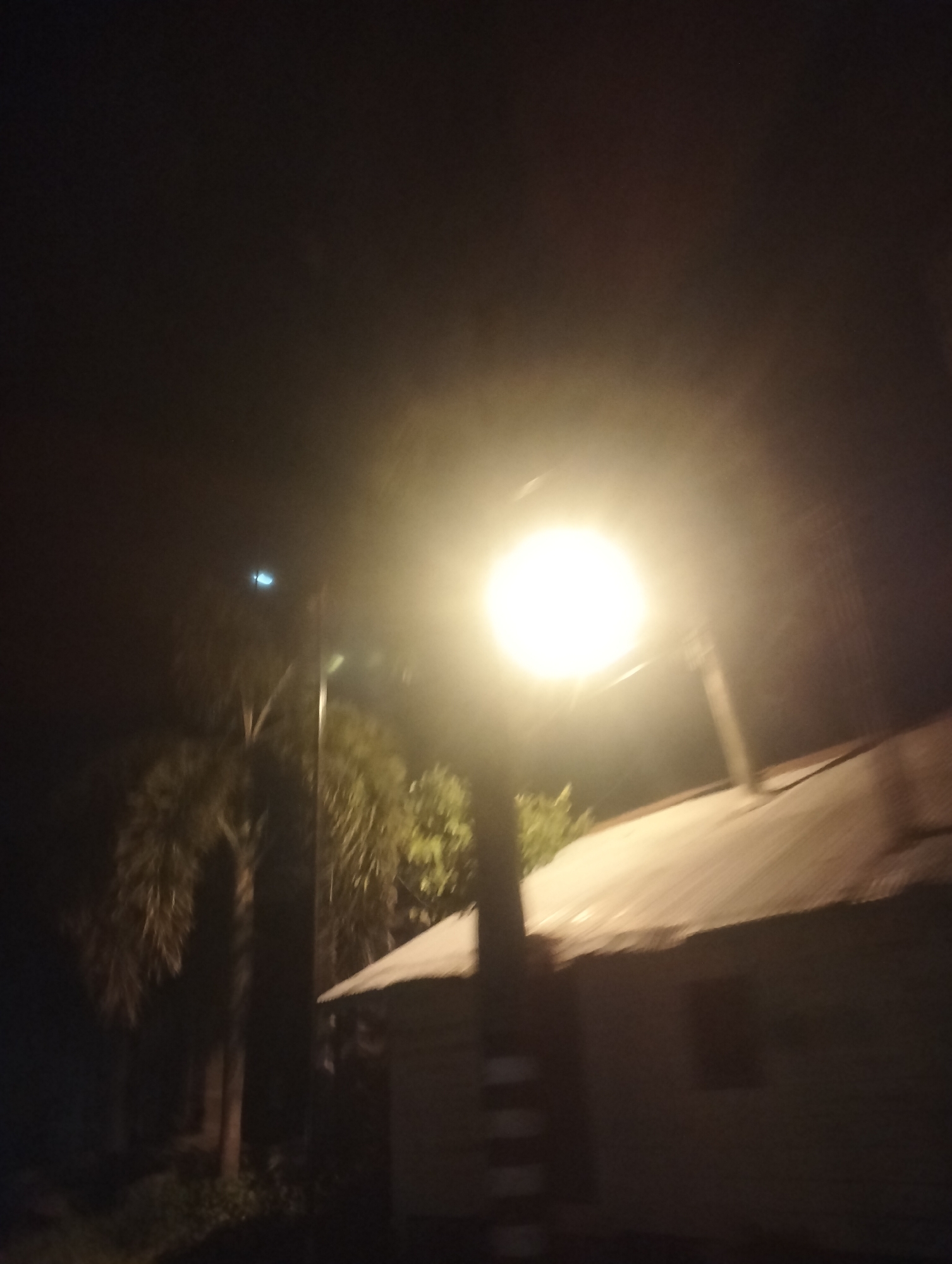 The image depicts a night scene in Lhokseumawe, Indonesia, illuminated by a bright streetlamp. The foreground is dominated by the intense glow of the lamp, which casts a wide, hazy light. To the right, the corrugated metal roof of a building is visible, appearing weathered and possibly aged. The building itself is dark and indistinct. To the left and behind the streetlamp, trees with lush, dark foliage are present, suggesting a tropical environment. A faint, blueish light is also visible in the sky above the trees, its origin unclear but potentially from another light source or reflection. The overall impression is of an outdoor area at night, with artificial lighting creating a stark contrast with the darkness.