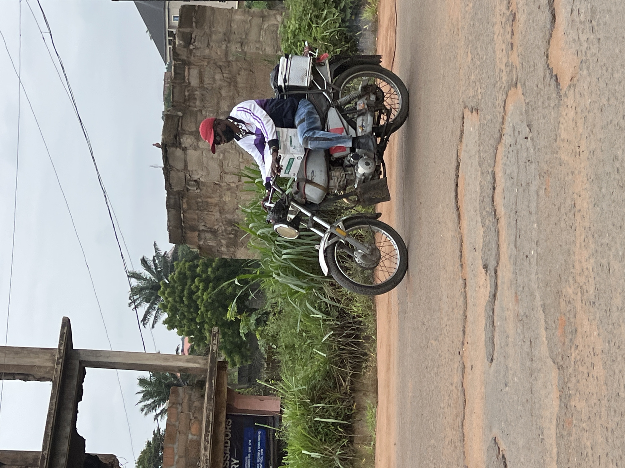 A man wearing a red cap, dark sunglasses, a white and purple jacket, and blue jeans is riding a motorcycle on a paved road with numerous potholes in Nkpor, Nigeria. He has a white box on the front of the motorcycle and a silver storage container on the back. The man is facing forward, and his hands are on the handlebars. In the background, there is a partially constructed building with exposed bricks and concrete, lush green vegetation, and a stone wall. The sky is overcast.