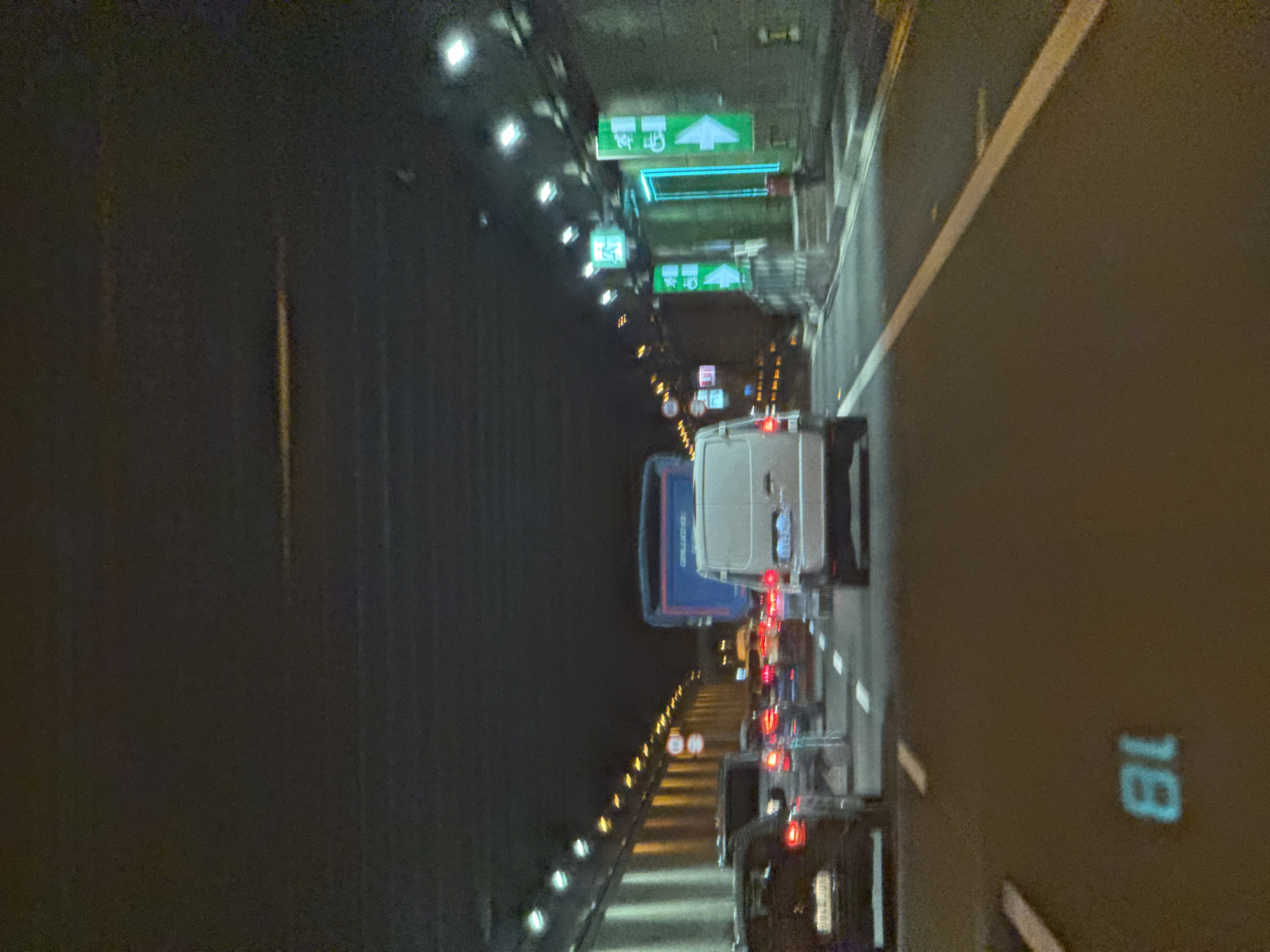 The image captures a night-time view from within a vehicle on a highway in Sant Joan d'Alacant, Spain. The scene is a traffic jam, with several vehicles visible ahead, including a white van and a dark-colored car. Red brake lights illuminate the darkness, indicating the slow-moving or stopped traffic. Overhead signs are illuminated with green and blue lights, providing directional information. One sign displays an arrow pointing upwards and to the right, accompanied by text that is difficult to decipher due to the image quality and angle. Another sign is predominantly blue. The road surface is dark, with a visible white lane marker. A pale yellow line runs along the side of the road, possibly a median or shoulder marking. The lighting suggests it is nighttime, and the weather appears clear. The overall mood is one of congestion and a typical urban commute at night. A number "18" is visible in a bright, illuminated blue on the road surface on the right side of the frame.