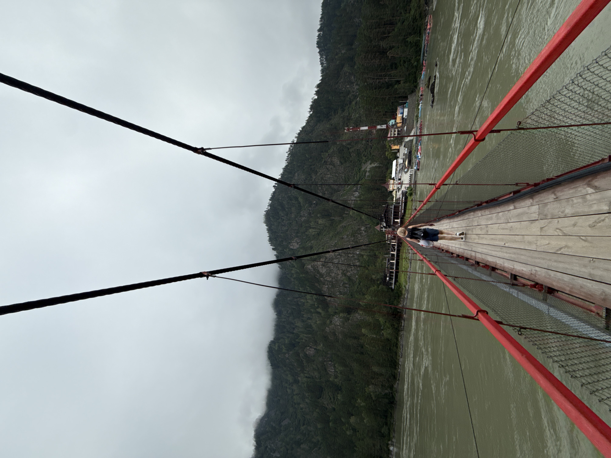 A lone figure walks across a wooden suspension bridge with red railings. The bridge spans a wide, choppy river with greenish-brown water. On the far bank, a settlement with several buildings, including a tall red and white tower, is nestled at the base of a steep, tree-covered mountain. The sky is overcast and grey. The scene appears to be the suspension bridge over the Katun River in the village of Elekmonar, near the border of the village of Barangol, Russia.