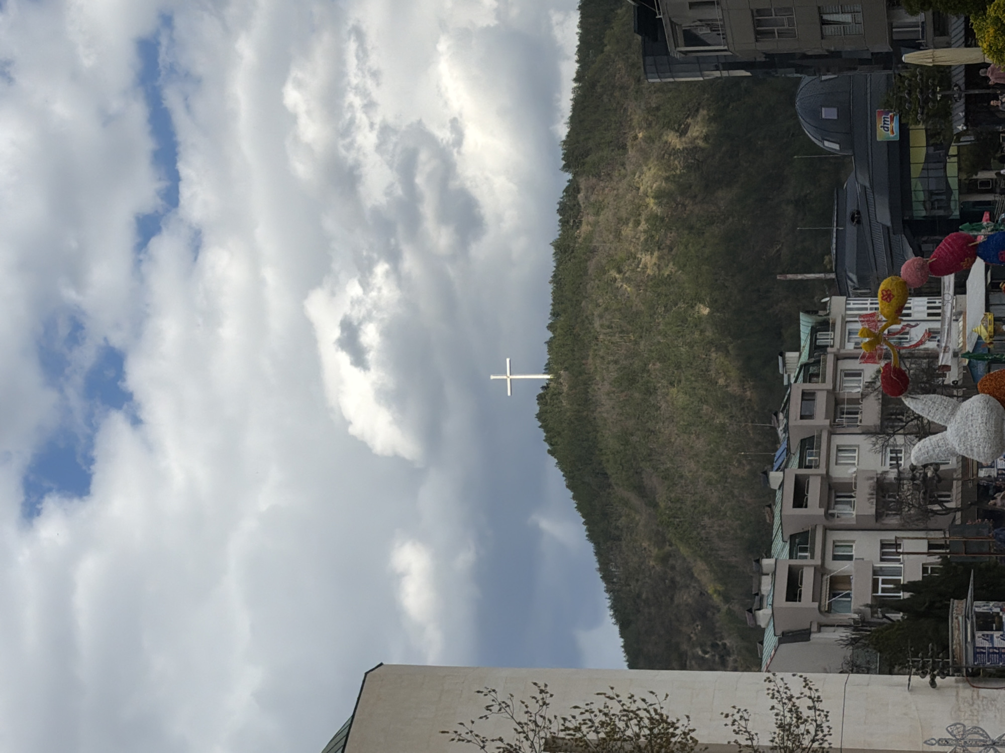 The image depicts a landscape in Blagoevgrad, Bulgaria, featuring a prominent hill covered in dense vegetation. A large white cross stands atop the hill, contrasting against the cloudy sky. In the foreground, a building with a light-colored facade is visible. The right side of the image shows a portion of the urban environment with buildings, including a pharmacy. A display of colorful decorations with easter bunnies adds some festive elements to the scene. People are strolling in the background. The sky is filled with grey and white clouds with glimpses of blue.