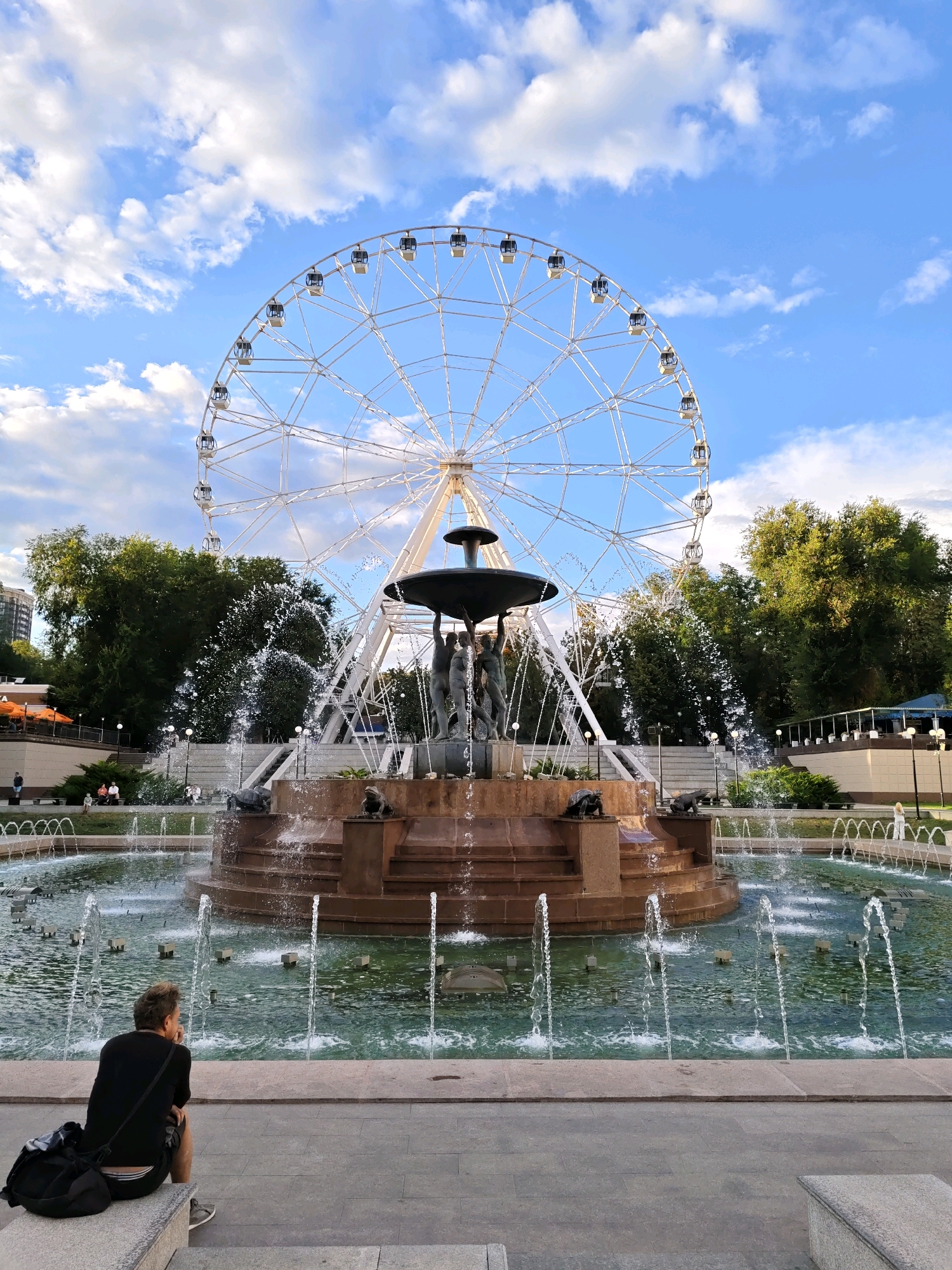 The image captures a scenic view of a Ferris wheel and a fountain in a park-like setting. The Ferris wheel, a prominent attraction, is large and white with numerous gondolas, set against a clear blue sky with scattered white clouds. Below the Ferris wheel, a multi-tiered fountain with sculpted figures and water jets creates a lively aquatic display. In the foreground, a person is seated on a stone bench, facing away from the camera, contemplating the view. The person is wearing a black t-shirt and shorts, with a black backpack placed beside them. The environment appears to be a public park with lush green trees and pathways, suggesting a recreational area. This scene is located at the Rostov Ferris Wheel (also known as the "Basta" or "Olimp-2") which is situated in the city of Rostov-on-Don, Russia.