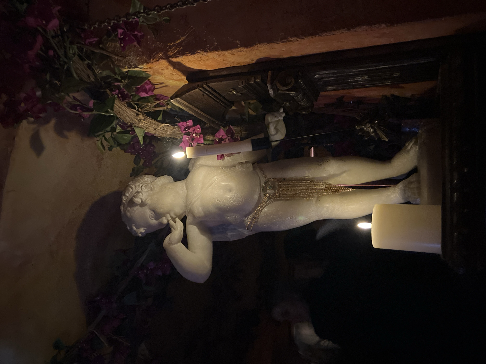 A sideways shot depicts a white marble or plaster statue of a reclining cherub or child, positioned near a mantelpiece decorated with purple flowers. The statue is illuminated by a single candle held in its hand, as well as another candle situated at its feet. The location is likely within an interior setting in Paris, France, possibly a vintage shop, a private residence, or a similar space with an antique or ornate aesthetic.