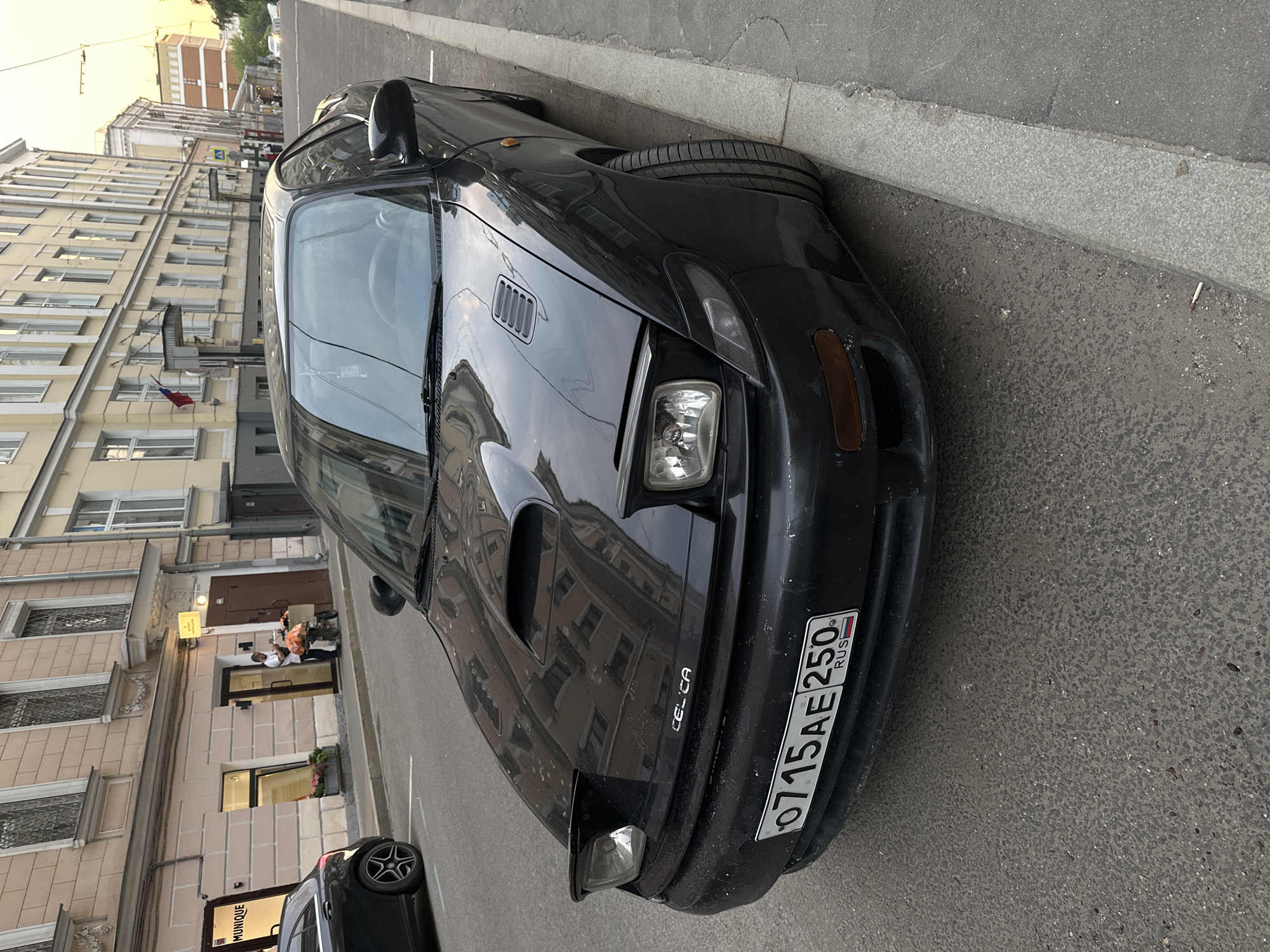 A black Toyota Celica is parked on a street in Moscow, Russia. The car is angled toward the curb on the right side of the frame. The license plate reads "O715AE250 RUS." The license plate is dirty. A building with multiple windows is visible in the background to the left of the car. The building is several stories tall and has a light-colored facade. Further down the street, a small group of people is sitting outside. A dark-colored car is partially visible in the lower left corner. The street appears to be paved and is relatively clean. The weather is overcast.