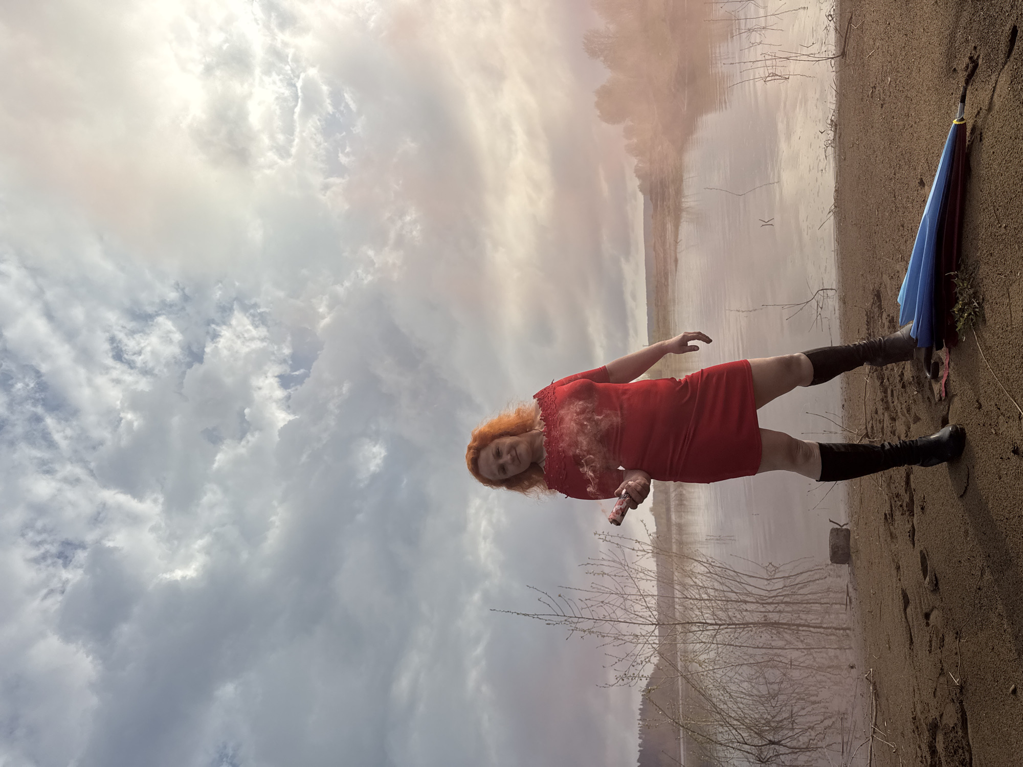 The image shows a red-haired woman in a red dress and black boots standing on a sandy shore. She is holding a smoking device that emits a pinkish cloud. Behind her is a body of water with bare trees along the opposite shore. The sky is cloudy and overcast. A blue and maroon umbrella lies on the sand to the right. The photo appears to have been taken with the camera tilted. The geographic location is not evident.