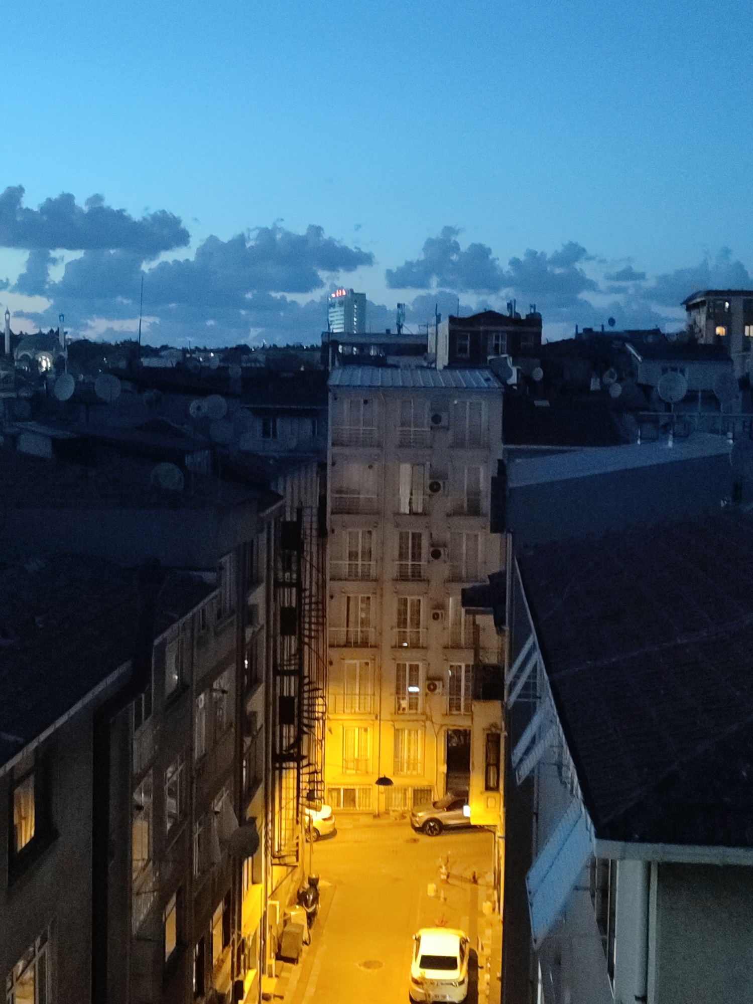 The image depicts an urban street scene at dusk in Kadıköy, Turkey. The sky is a deep blue with scattered dark clouds. The foreground is dominated by the facades of multi-story buildings lining a narrow street. The buildings are a mix of styles, with some featuring balconies and air conditioning units. Streetlights cast a warm yellow glow on the pavement and the lower levels of the buildings.
Several vehicles are visible in the street. A white hatchback is parked towards the bottom center of the frame, and a silver SUV is parked further up the street. There appear to be no people actively engaged in any discernible actions. The overall impression is one of a quiet evening in a residential or mixed-use urban area.
