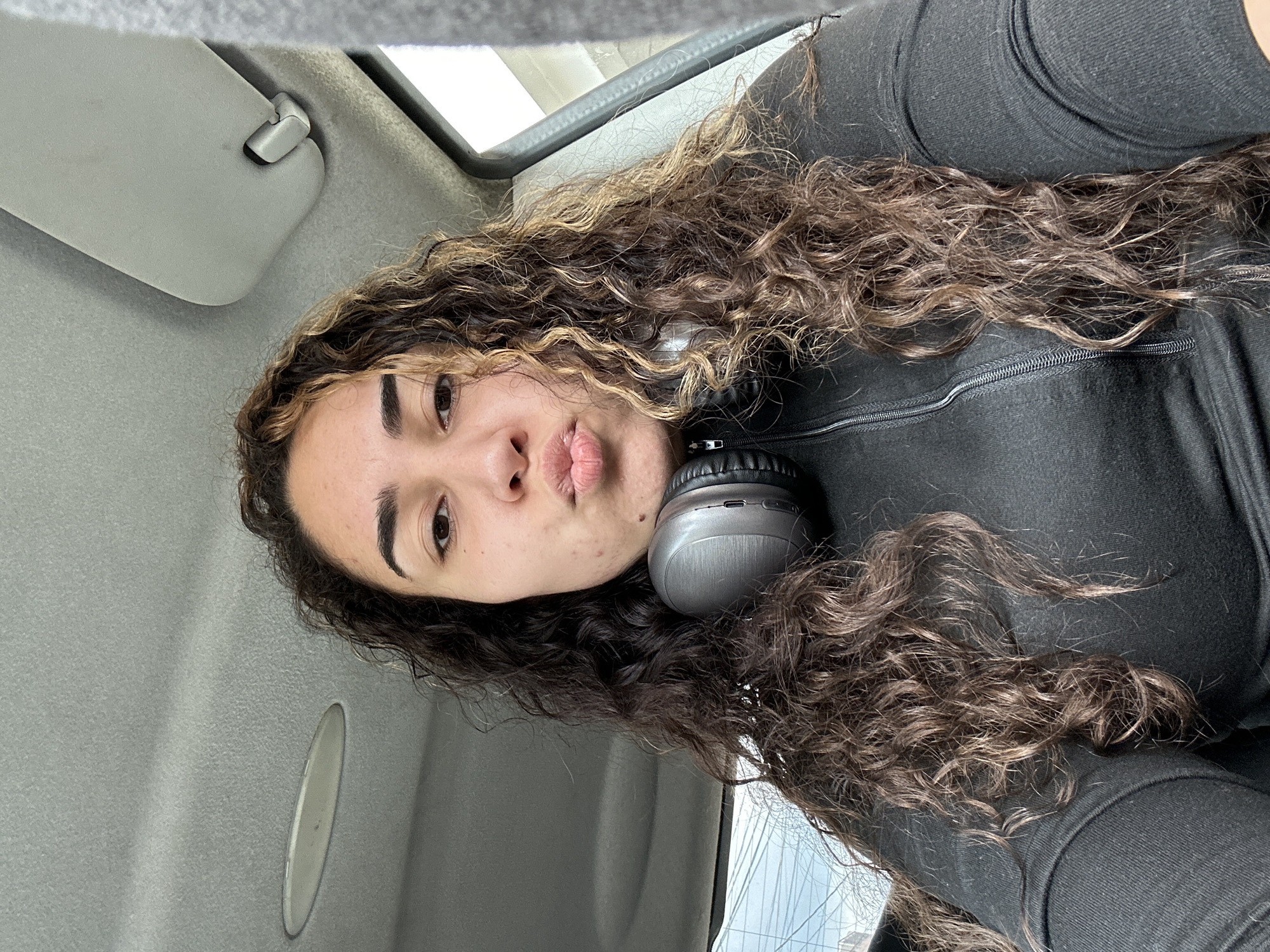 The image is a selfie taken inside a car. The subject is a person with curly brown and blonde hair, wearing a black zippered top and headphones around their neck. The interior of the car is visible, including the grey ceiling and a visor. Outside the window, there's a glimpse of sky and what appears to be power lines. The location cannot be definitively identified as Burgas, Bulgaria, based on the provided visual information.