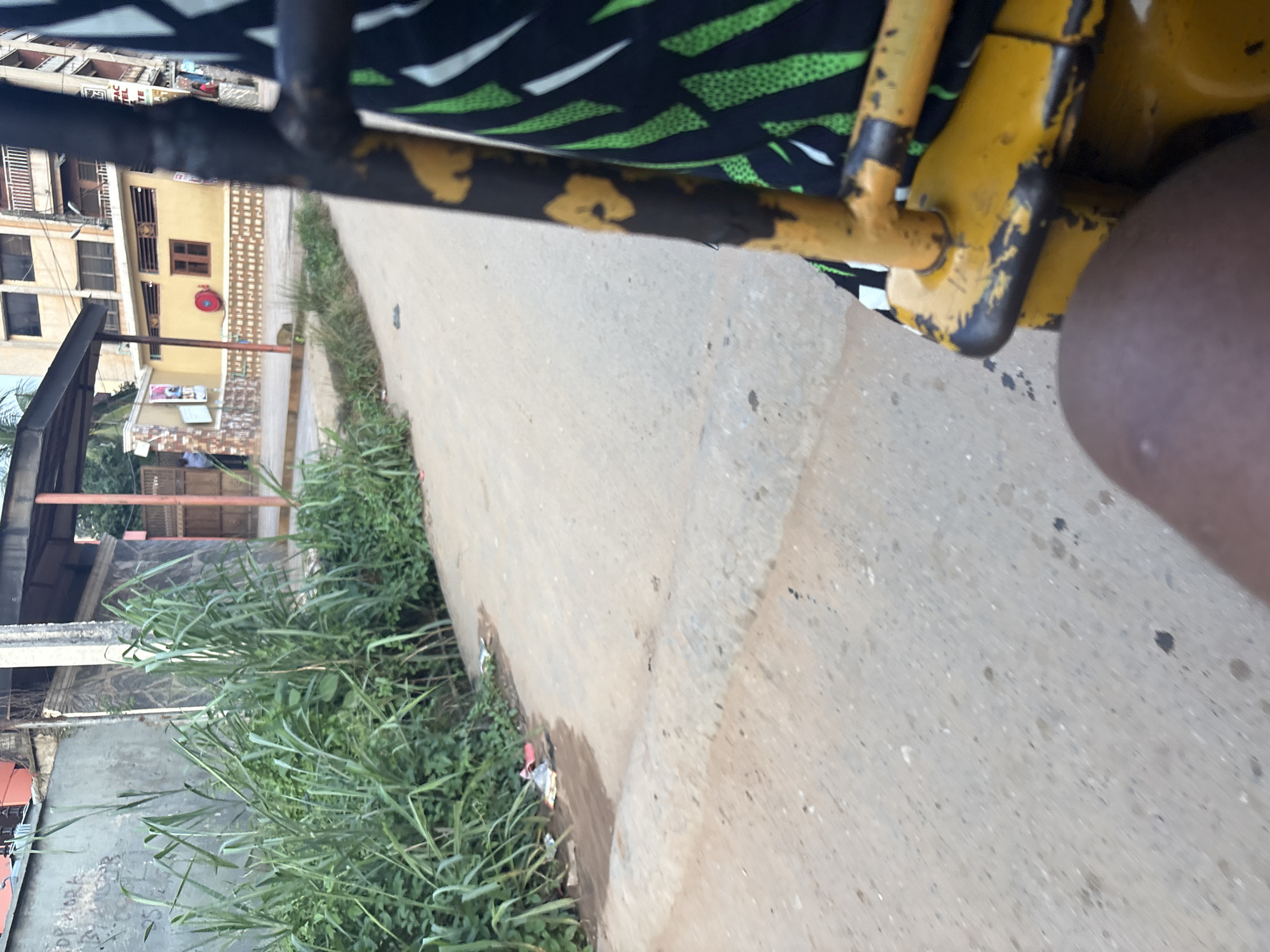 A person is seated in a yellow, metal-framed vehicle, possibly a tricycle or motorcycle taxi, in Onitsha, Nigeria. The vehicle's frame is worn and has chipped yellow paint, revealing black underneath. The passenger is wearing a black and green patterned garment. The vehicle is on a paved, grey road with some dark stains and debris. To the left, a wall with graffiti and overgrown green vegetation lines the road. Beyond the vegetation, a building with an orange facade and a covered walkway is visible. Further down the road, a light yellow building with a red fire extinguisher mounted on its wall and a small wooden-framed window stands. The sky is not visible in the frame. The scene appears to be in an urban environment with a mix of residential and commercial structures.