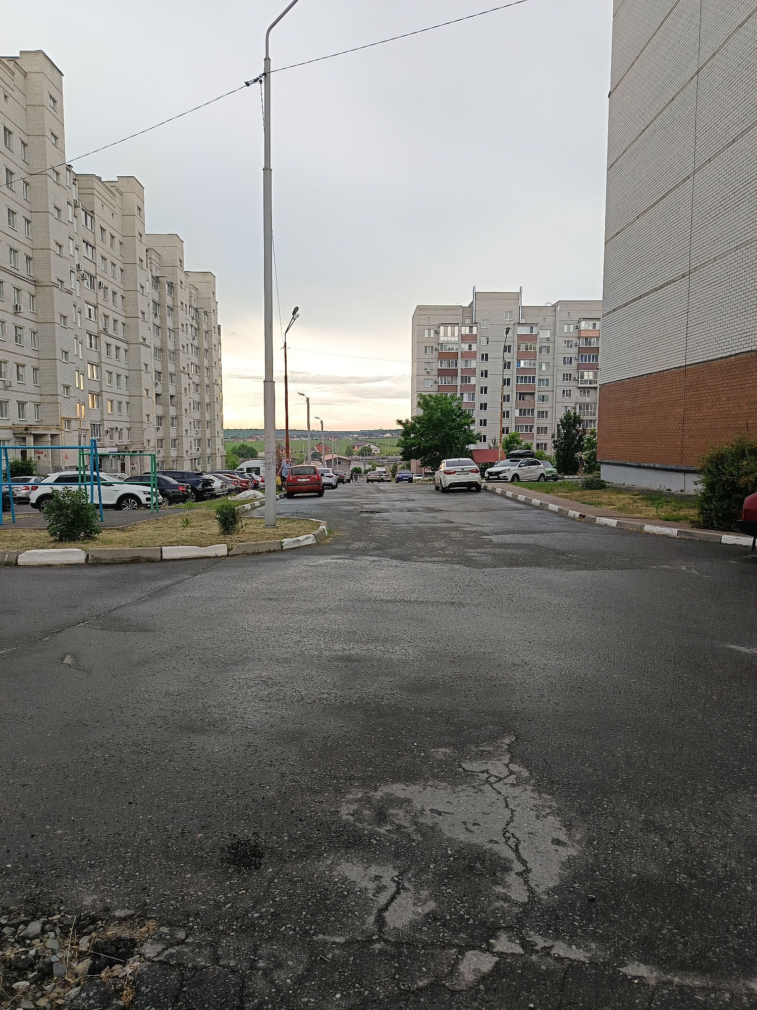 The image depicts an overcast day in a residential area of Gubkin, Russia, likely a courtyard between apartment buildings. The foreground is dominated by a wet asphalt road with visible cracks and patches. To the left, a multi-story apartment building, constructed with white bricks, stretches along the side of the frame. In front of this building, a row of parked cars is visible, including a white SUV, a black sedan, and other vehicles. A small patch of grass and a playground structure with blue and yellow bars are also present. A prominent lamppost stands near the center of the image, with power lines extending across the frame.
The middle ground reveals another apartment building on the right, also clad in white brick with sections of red brick near the base. More parked cars line the street, including a white sedan, a green car, and a red sedan further down. A few trees and bushes are scattered throughout the scene.
In the background, more apartment buildings of similar architectural style can be seen. The sky is cloudy and grey, with a hint of soft light on the horizon suggesting either early morning or late evening. The overall impression is one of a typical urban residential environment.
