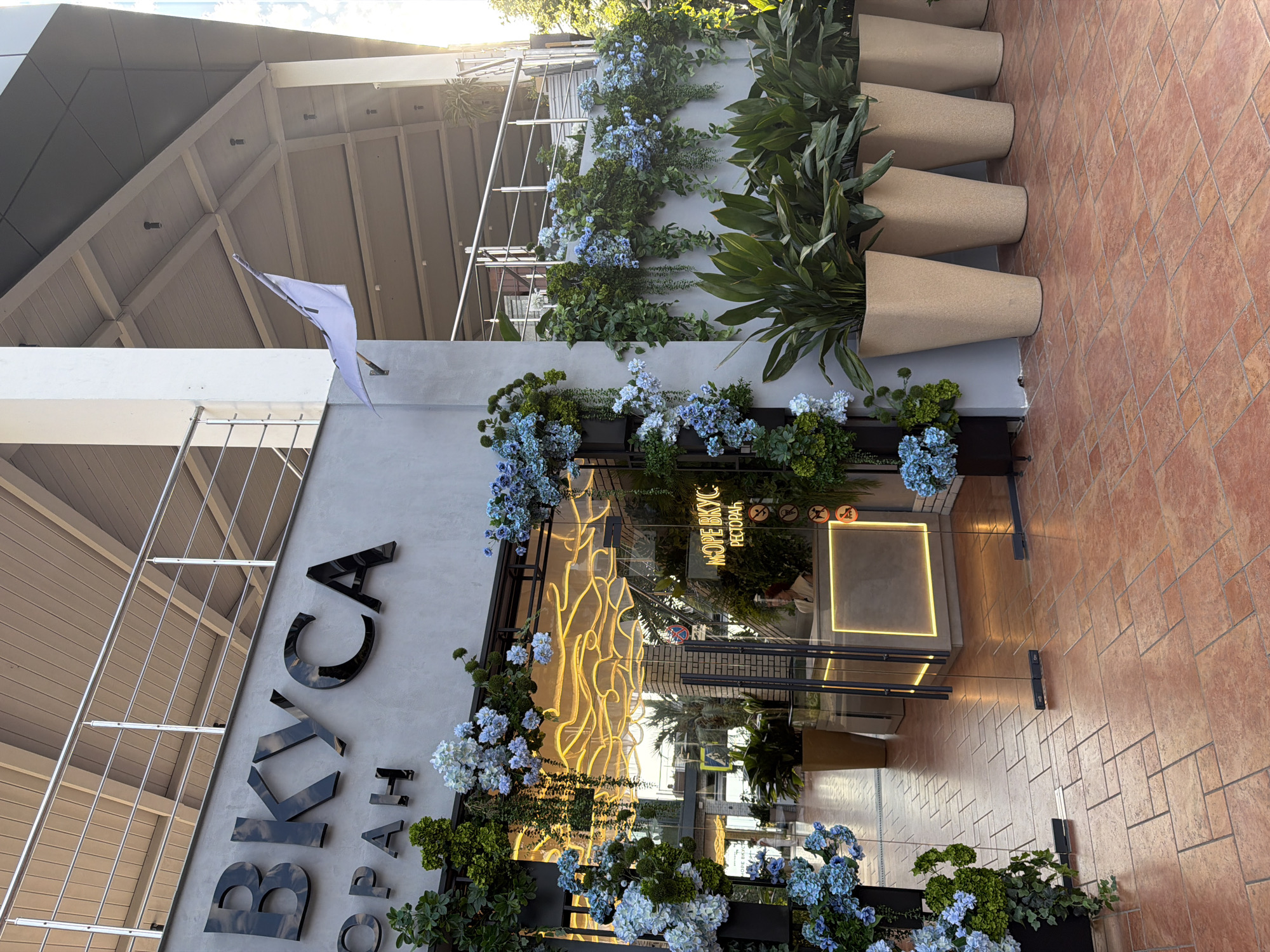 This is a medium shot of the exterior of a restaurant in Sochi, Russia. The restaurant has a modern design with a gray facade and large glass doors. Above the entrance, the word "ВКУСА" is prominently displayed in large, black, three-dimensional lettering. Below this, smaller lettering reads "ОРАН".
The entrance is decorated with an abundance of artificial blue hydrangeas and green foliage, creating a lush and vibrant display. Inside the restaurant, a neon sign on the wall glows with yellow wavy lines. A reception desk is visible, with a woman in a light-colored blouse standing behind it.
Several large, light-colored planters with tall, leafy green plants are arranged in front of the facade, adding to the natural aesthetic. The ground is paved with reddish-brown tiles laid in a geometric pattern. A white flag with a blue emblem is mounted on the wall to the right of the main signage. The overall impression is that of a well-maintained and inviting establishment, likely situated in a public or commercial area of Sochi.