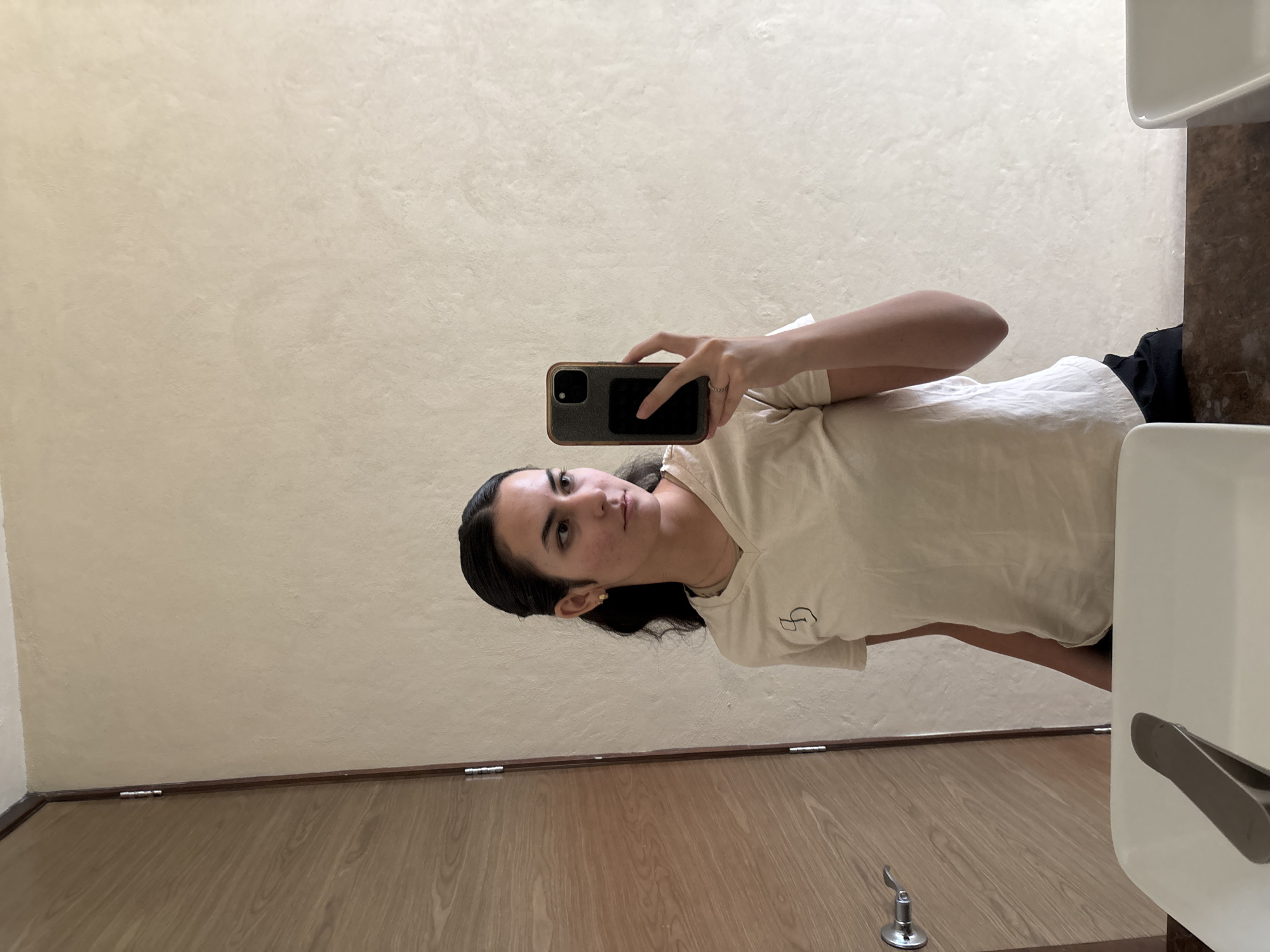 A young woman is taking a selfie with her smartphone in what appears to be a restroom. She is wearing a light-colored T-shirt and dark bottoms. The restroom features a light-colored wall, a wooden door, a sink, and potentially a hand dryer. The location is San Bernardino Tlaxcalancingo, Mexico.