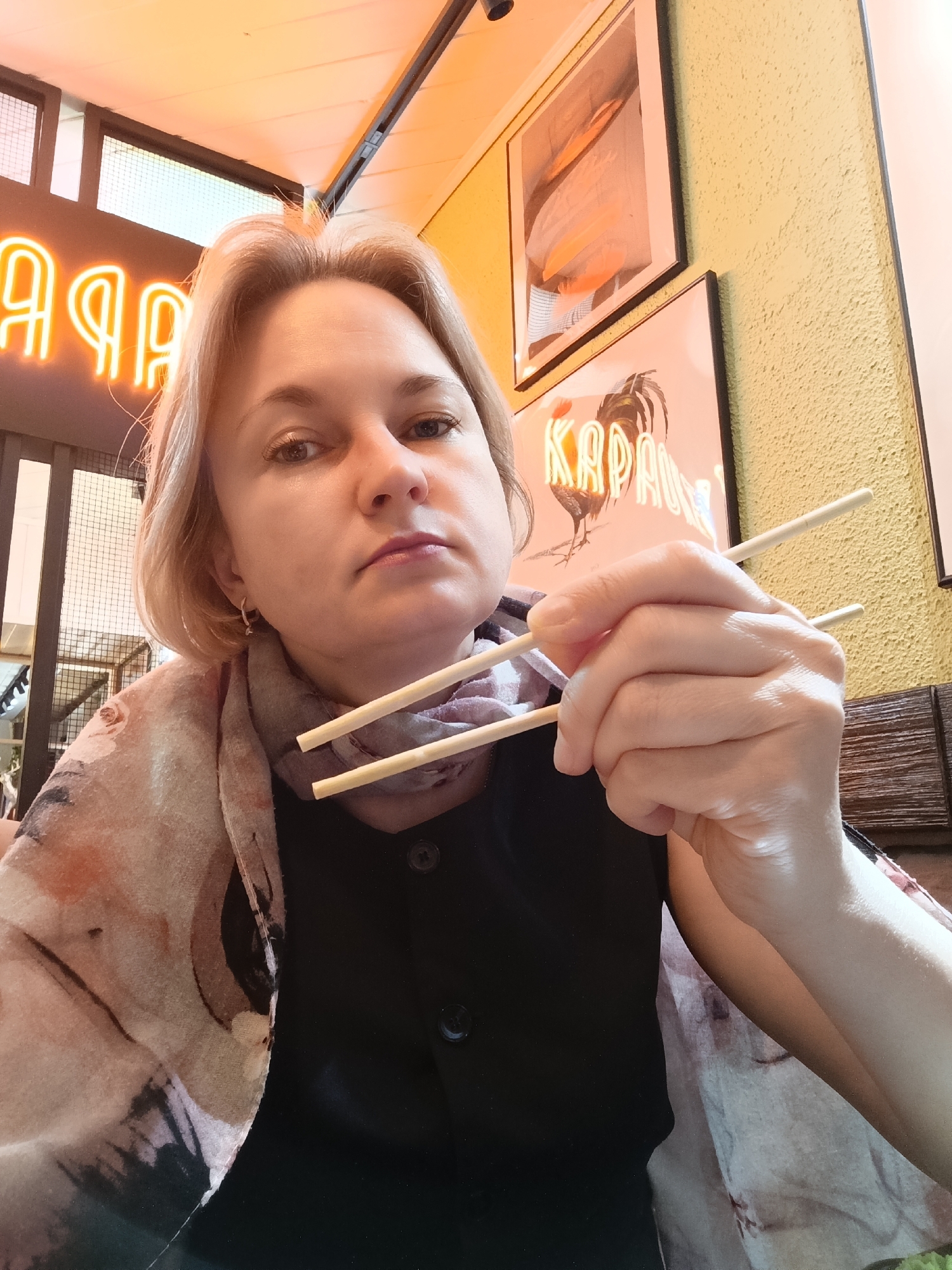 A close-up, eye-level portrait captures a blonde-haired woman indoors, likely within a restaurant or casual dining establishment in Chaikovsky, Russia. She wears a black top and a light-colored, patterned scarf around her neck, and gazes directly at the viewer with a neutral, calm expression. Her right hand is prominently featured, holding a pair of light wooden chopsticks as if preparing to eat.
The background is illuminated by warm, amber lighting, suggesting an indoor setting potentially during evening hours or a deliberately cozy ambiance. To the woman's upper left, a bright orange neon sign displays Cyrillic characters, appearing to read "ЯЛА" (YALA) from this perspective. To her upper right, two framed pictures are visible against a textured, light yellow wall. The lower picture clearly shows an illustration of a rooster and the Cyrillic word "КАРАБАШ" (KARABASH), possibly indicating a restaurant theme or name. The upper picture is less distinct. The overall scene conveys a moment of dining in a modern, casual eatery with specific cultural cues from the signage.